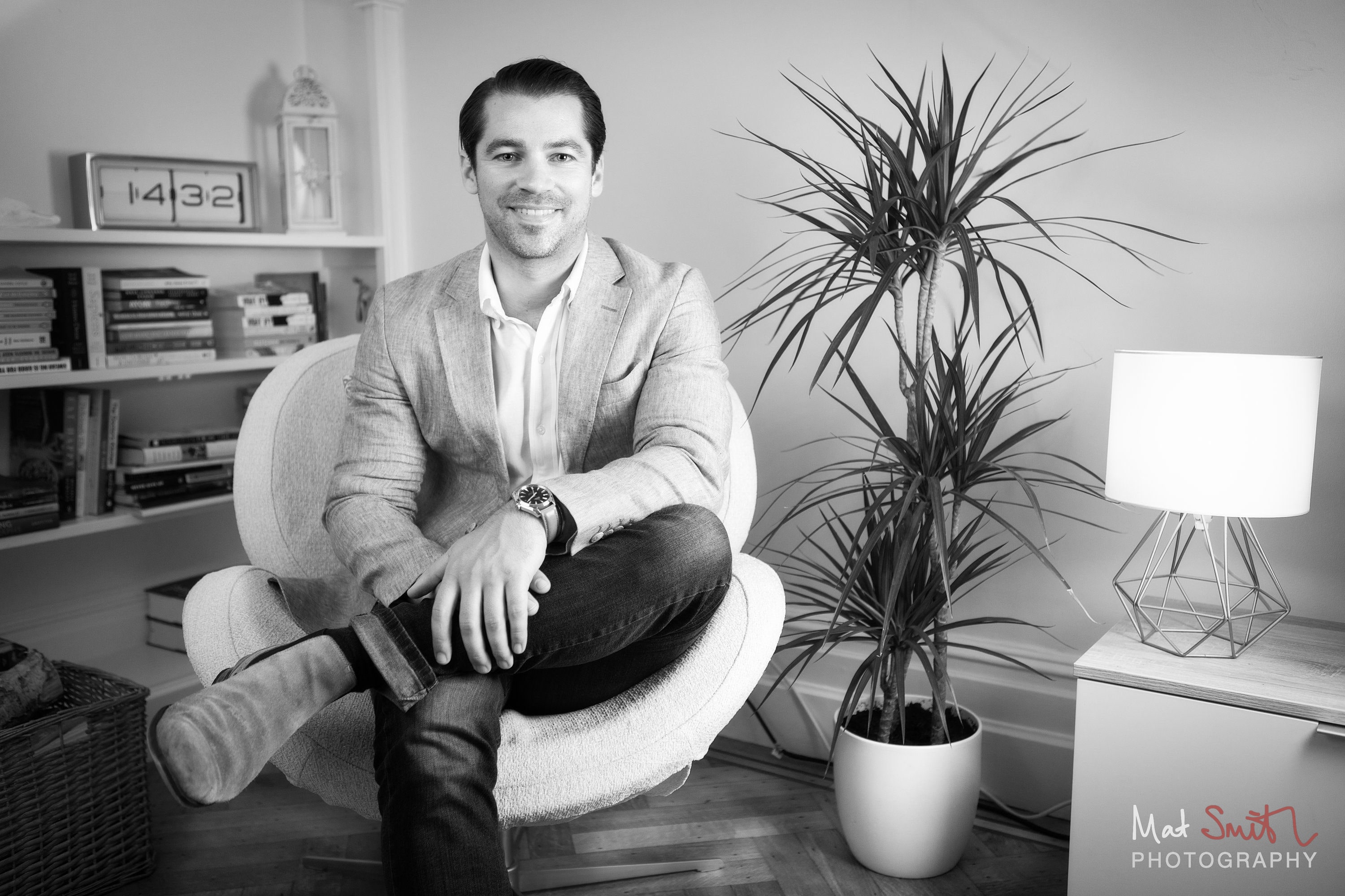 Black and white portrait of a man in a blazer sitting cross legged in an armchair beside a plant and bookshelves, portrait by Mat Smith Photography, London