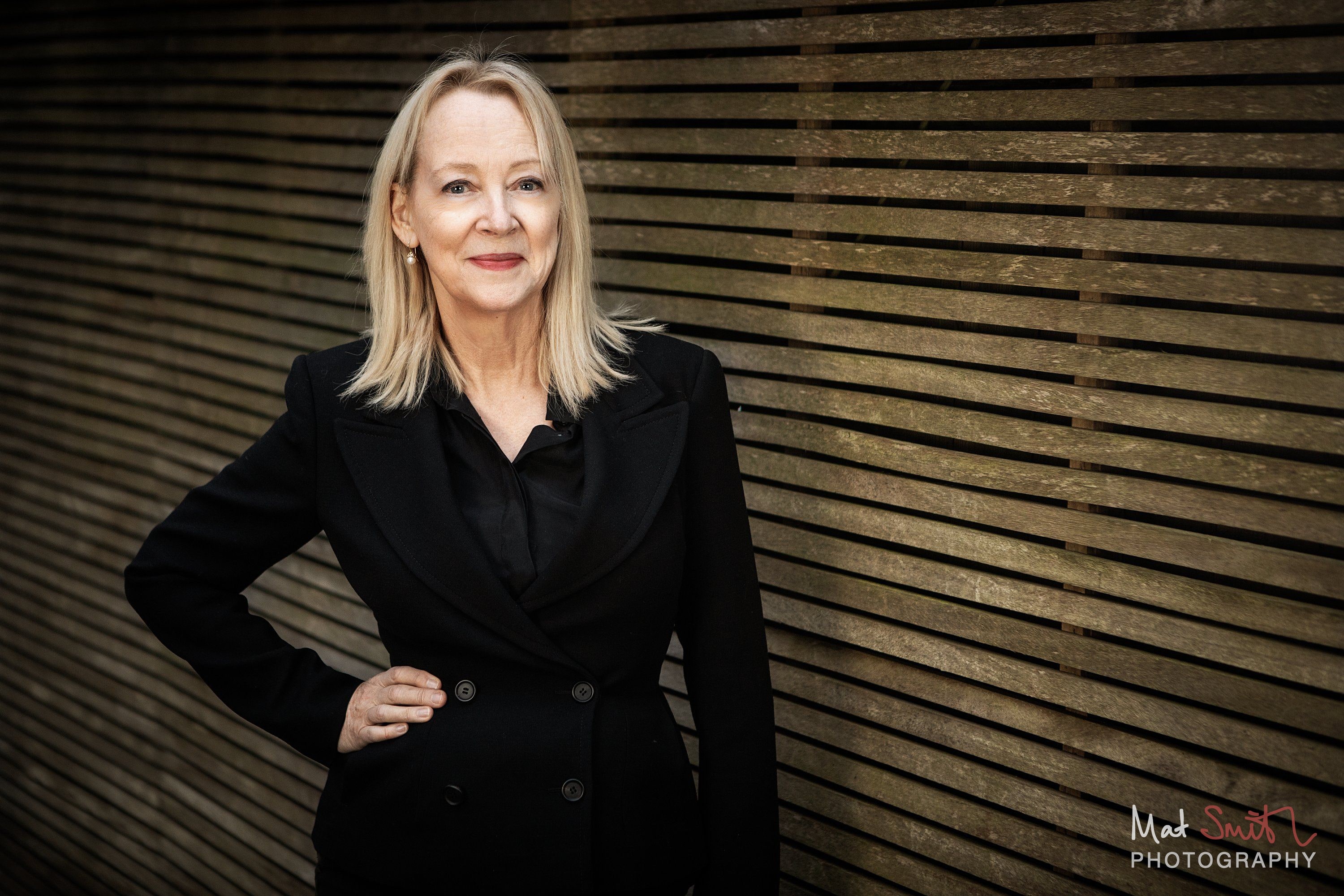 A woman with shoulder-length blonde hair in a black jacket stands with one hand on her hip against a slatted wooden garden fence, portrait by Mat Smith Photography, London
