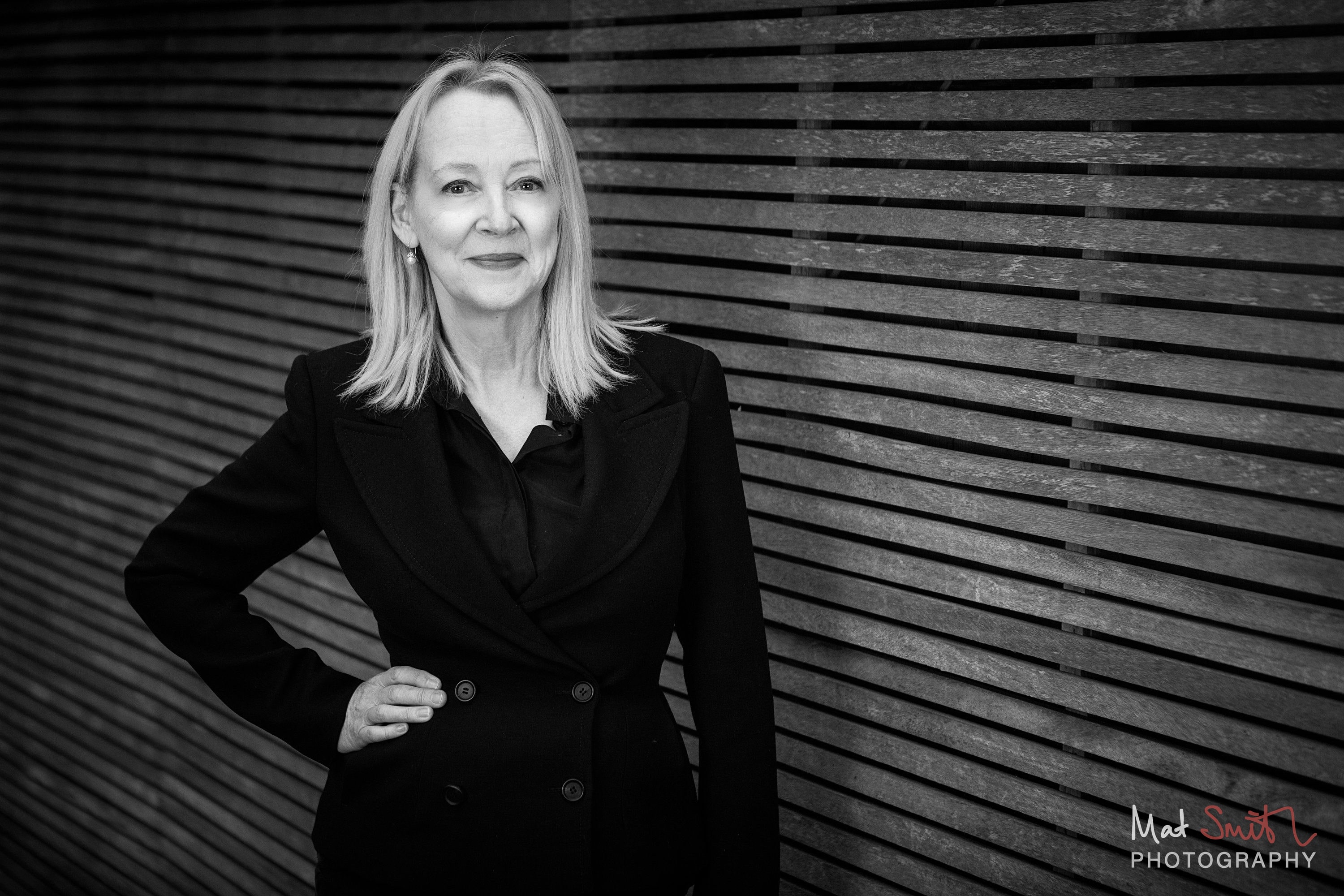 Black and white version of a woman with blonde hair in a black jacket, hand on hip, against a slatted wooden fence, portrait by Mat Smith Photography, London