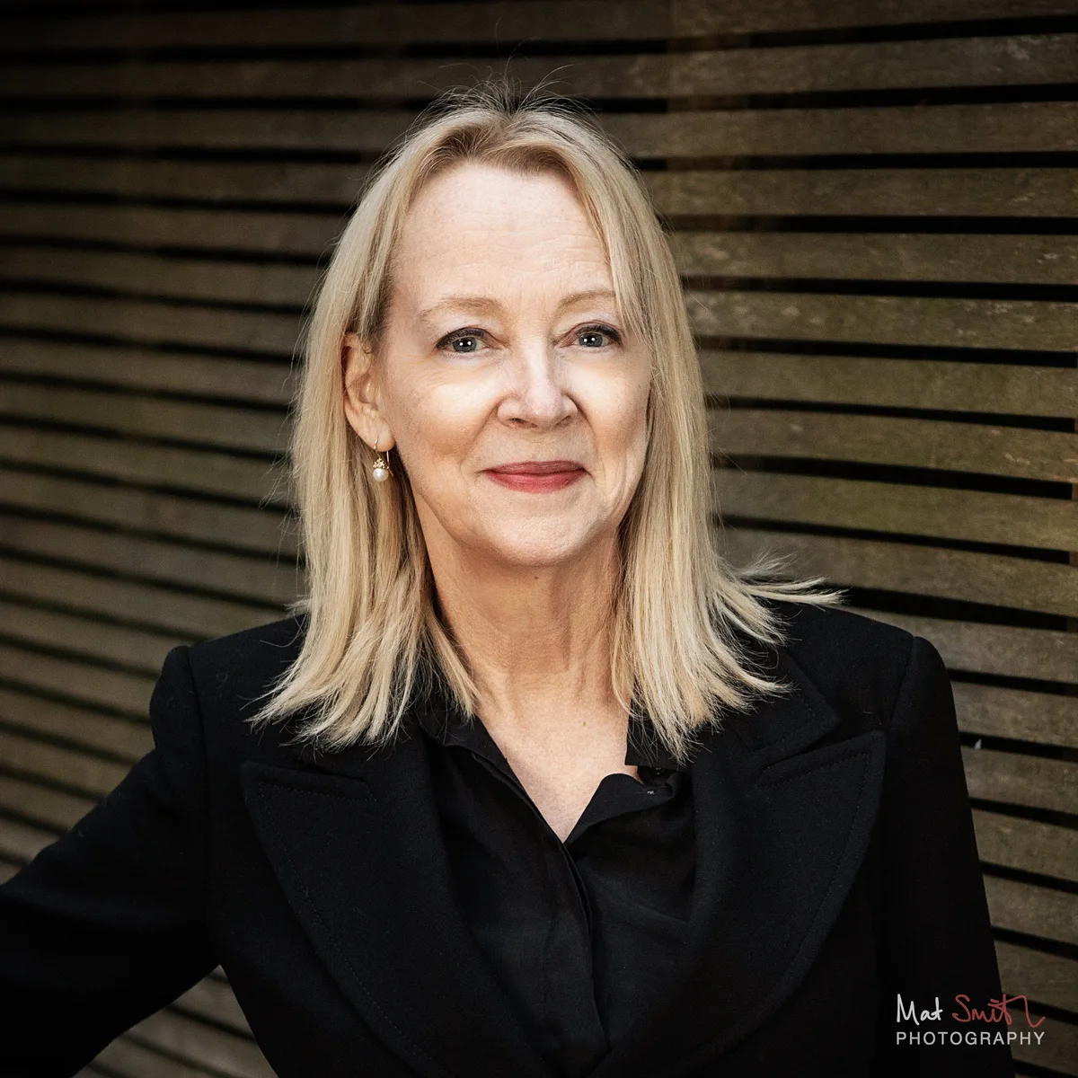 Close portrait of a smiling woman with blonde hair and pearl earrings in a black jacket in front of a slatted wooden fence, portrait by Mat Smith Photography, London