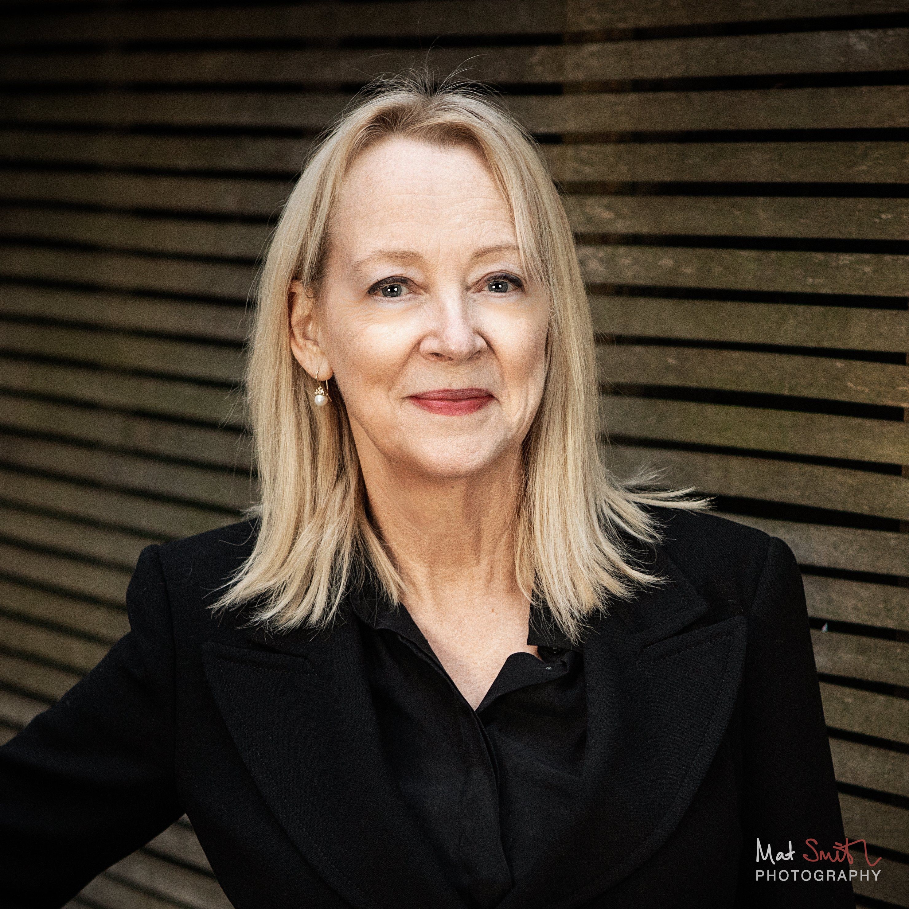 Close portrait of a smiling woman with blonde hair and pearl earrings in a black jacket in front of a slatted wooden fence, portrait by Mat Smith Photography, London