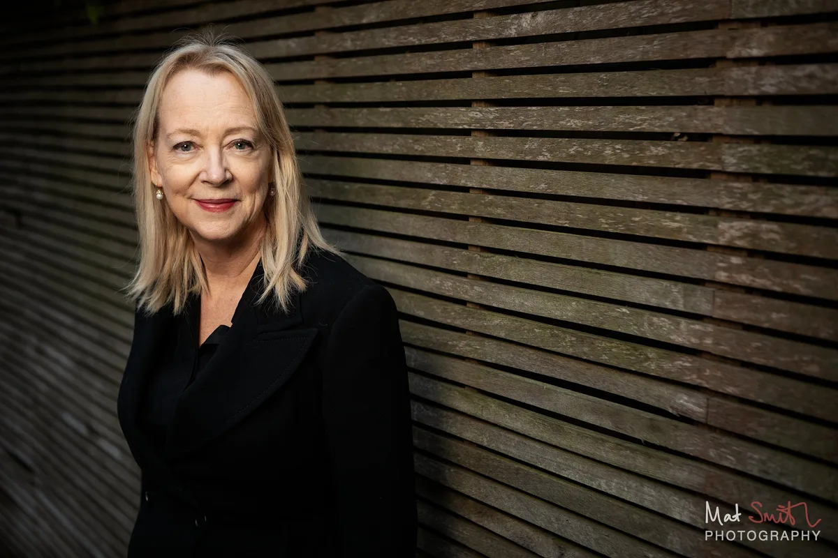 A woman with blonde hair in a black jacket smiles over her shoulder beside a slatted wooden garden fence, portrait by Mat Smith Photography, London