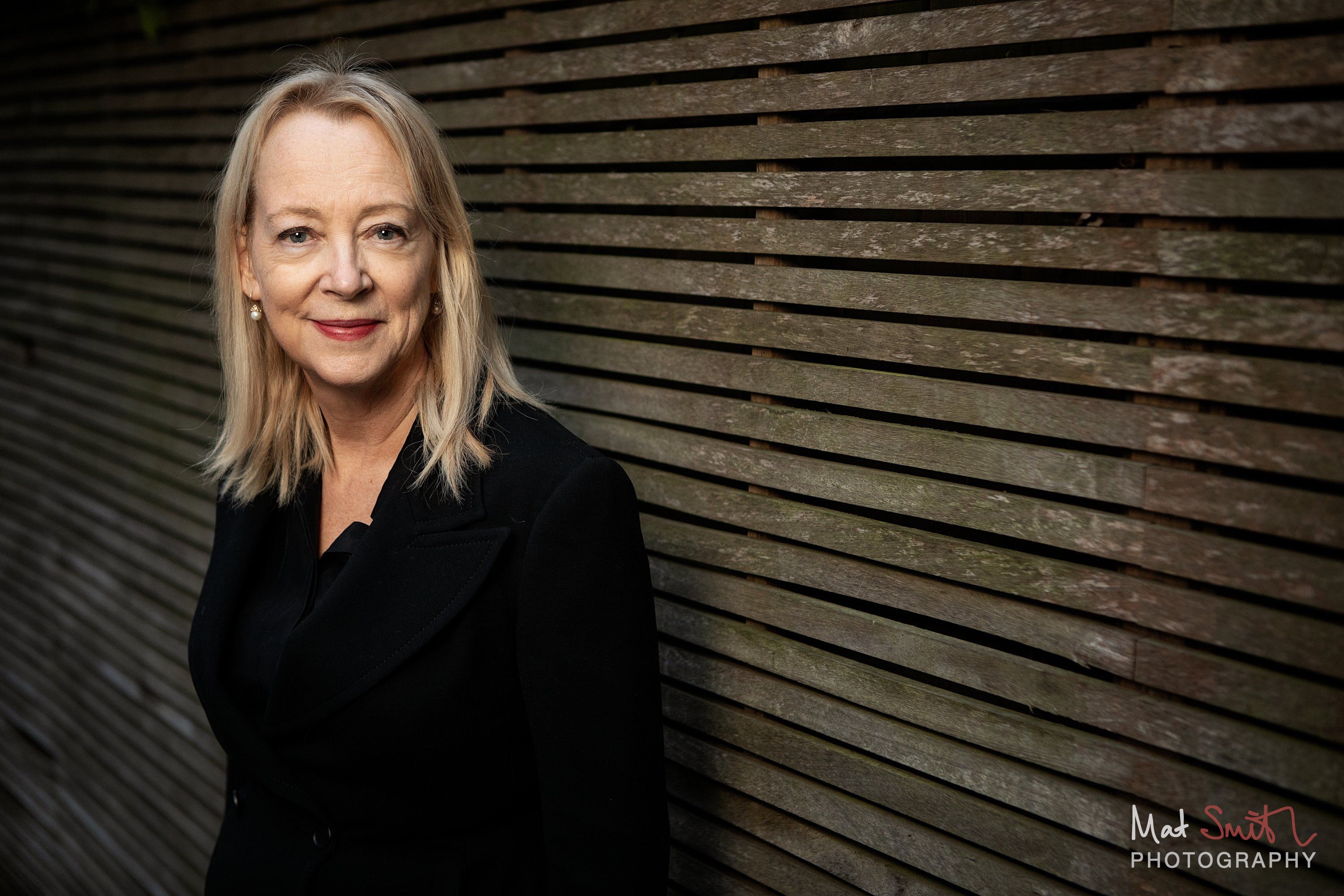 A woman with blonde hair in a black jacket smiles over her shoulder beside a slatted wooden garden fence, portrait by Mat Smith Photography, London