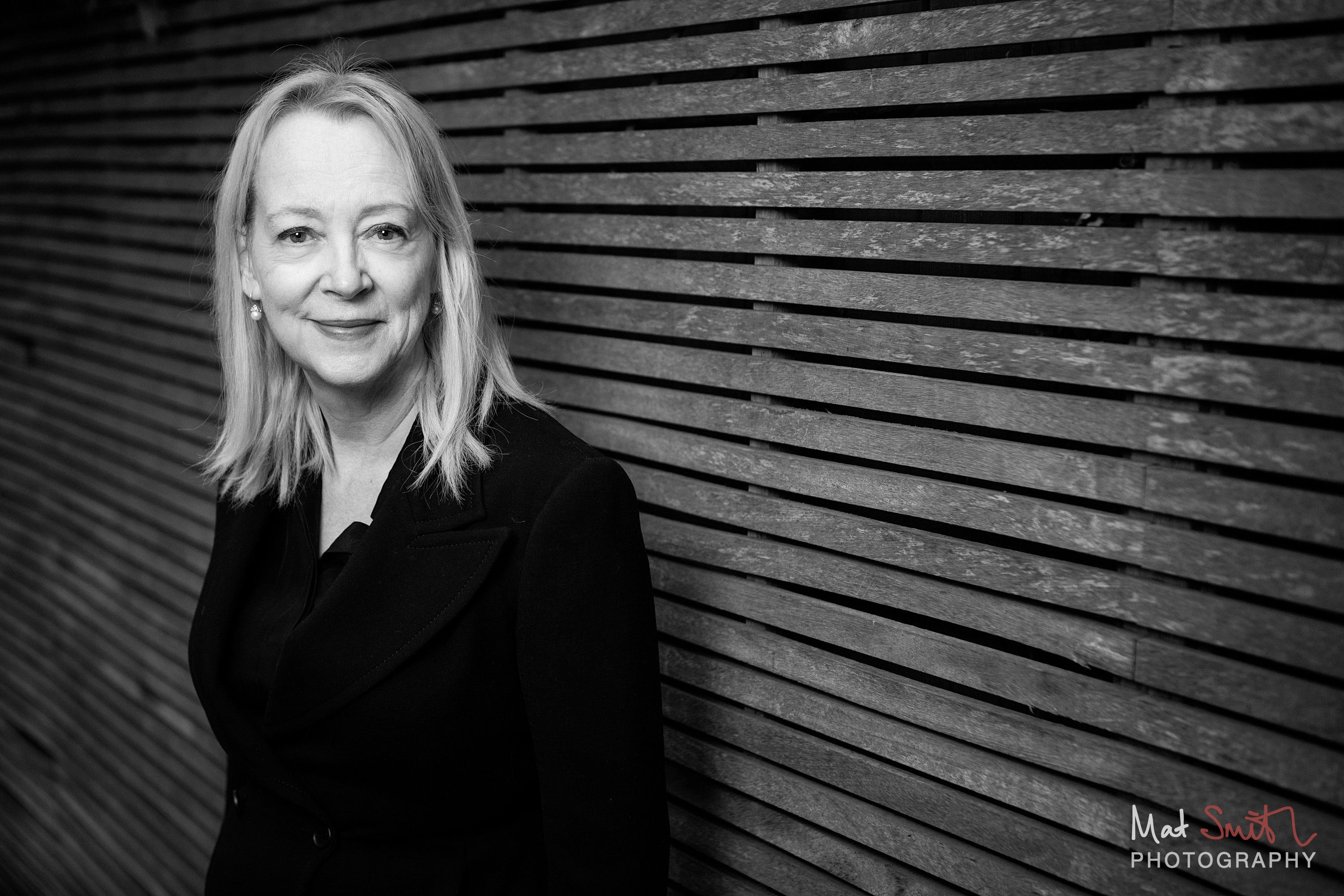 Black and white portrait of a woman with blonde hair in a black jacket smiling beside a slatted wooden fence, portrait by Mat Smith Photography, London