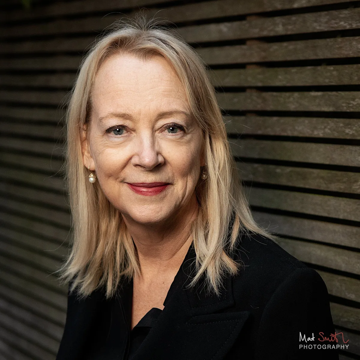 Tight headshot of a woman with blonde hair, pearl earrings and red lipstick smiling warmly against dark wooden slats, portrait by Mat Smith Photography, London