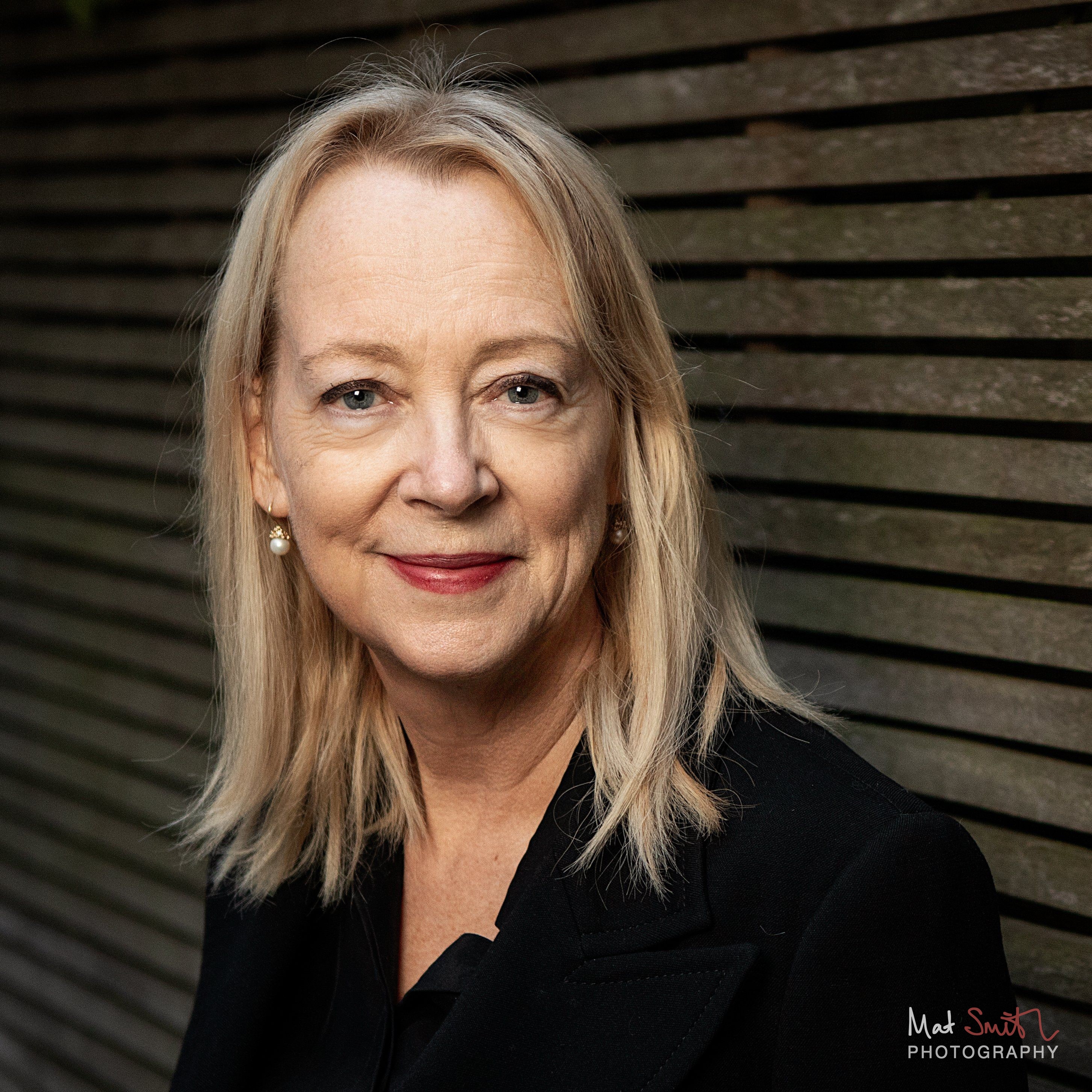 Tight headshot of a woman with blonde hair, pearl earrings and red lipstick smiling warmly against dark wooden slats, portrait by Mat Smith Photography, London