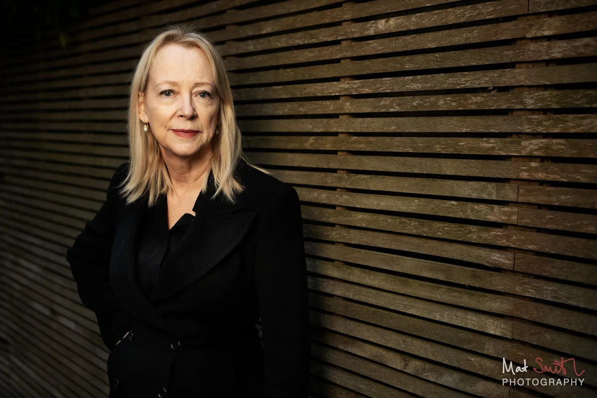 A woman with blonde hair in a black jacket looks calmly at the camera in front of a slatted wooden fence, portrait by Mat Smith Photography, London