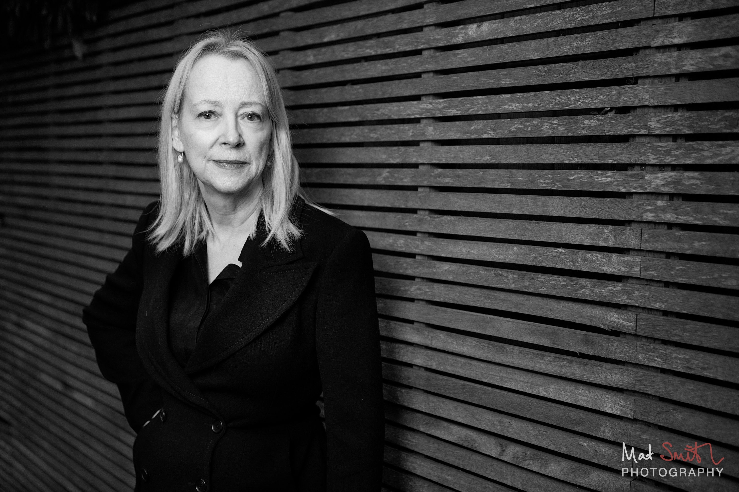 Black and white portrait of a woman with blonde hair in a black jacket with a calm expression against wooden slats, portrait by Mat Smith Photography, London