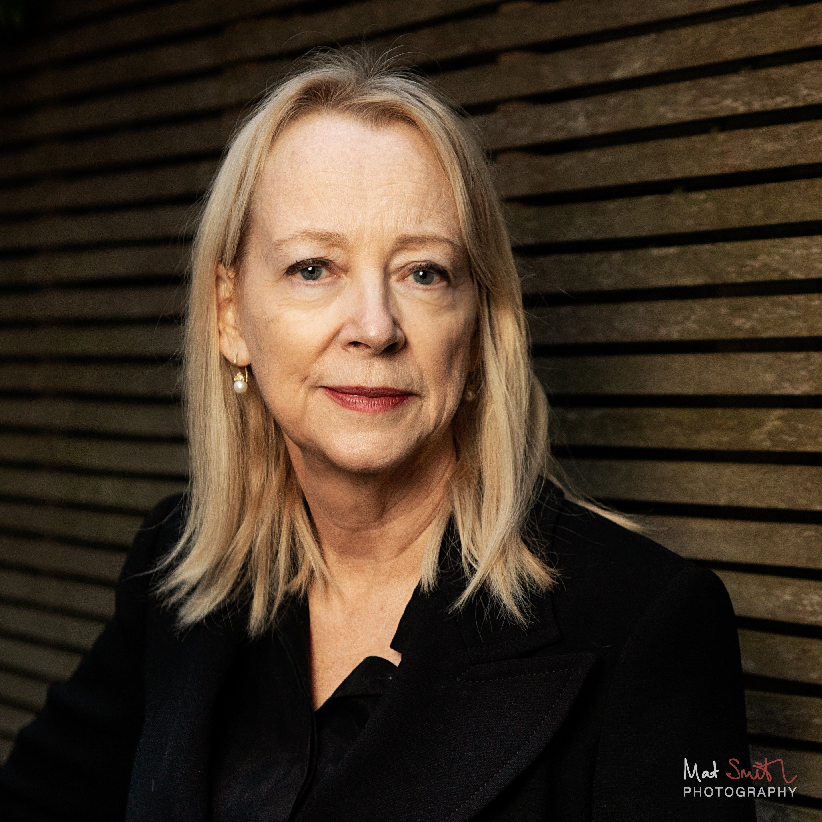 Headshot of a woman with blonde hair in a black jacket with a direct, composed gaze against a dark slatted fence, portrait by Mat Smith Photography, London