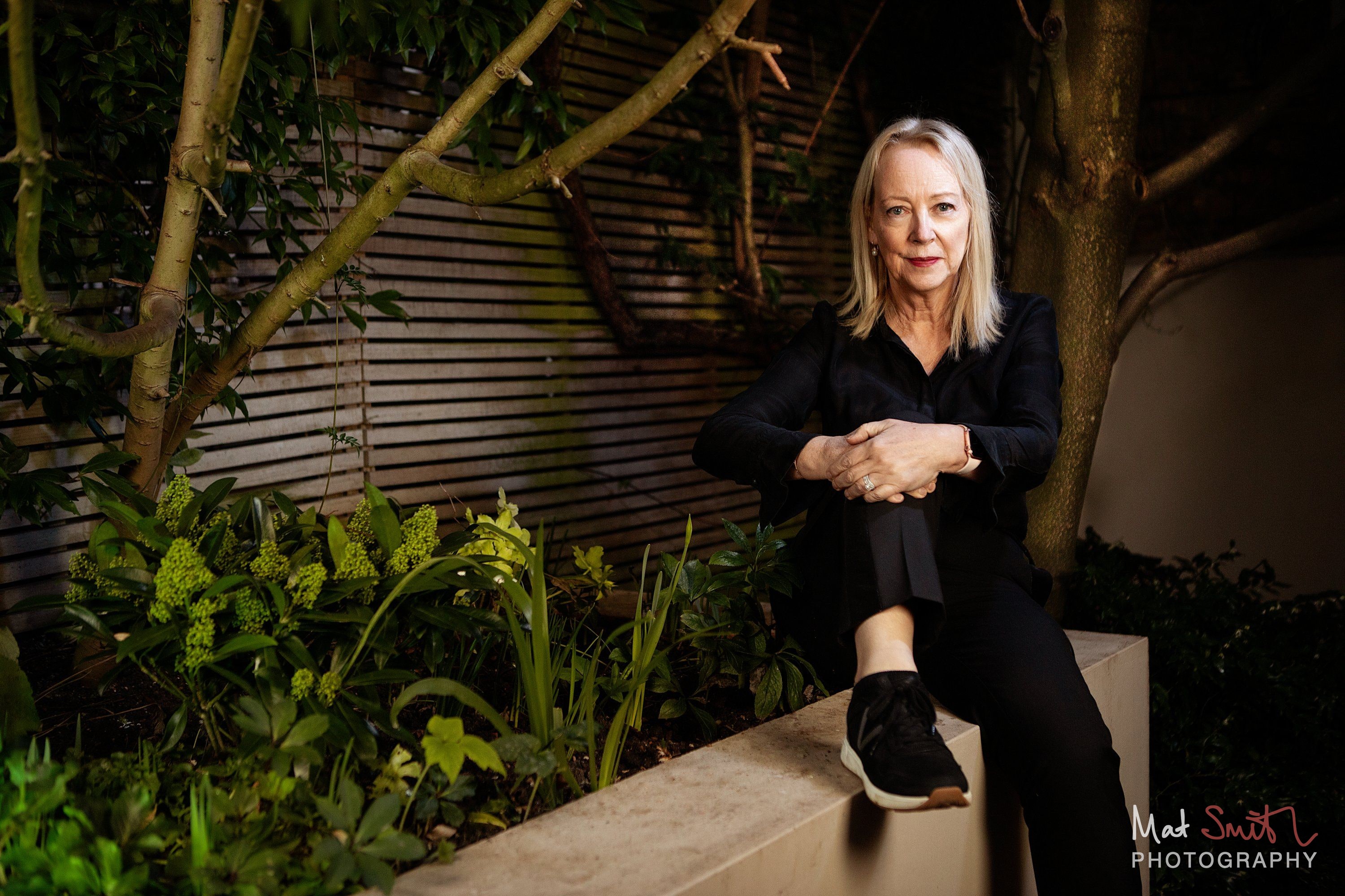 A woman in black sits on a low garden wall with her hands clasped over one knee, framed by branches and planting at dusk, portrait by Mat Smith Photography, London