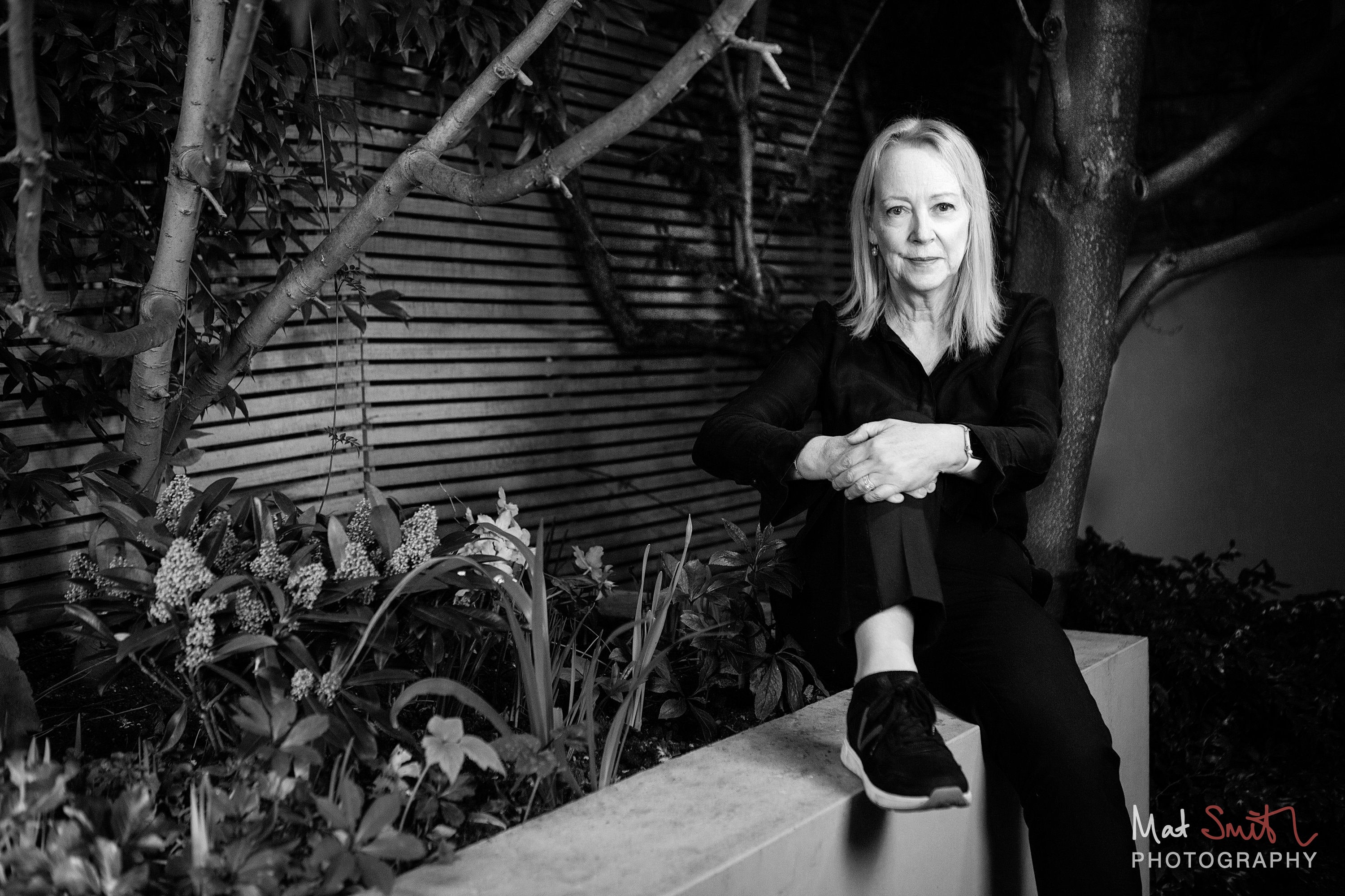 Black and white view of a woman in black seated on a garden wall among branches and plants, hands clasped over her knee, portrait by Mat Smith Photography, London