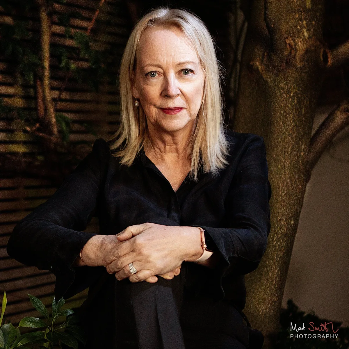 A woman with blonde hair in a black shirt leans forward with clasped hands beside a tree trunk in an evening garden, portrait by Mat Smith Photography, London