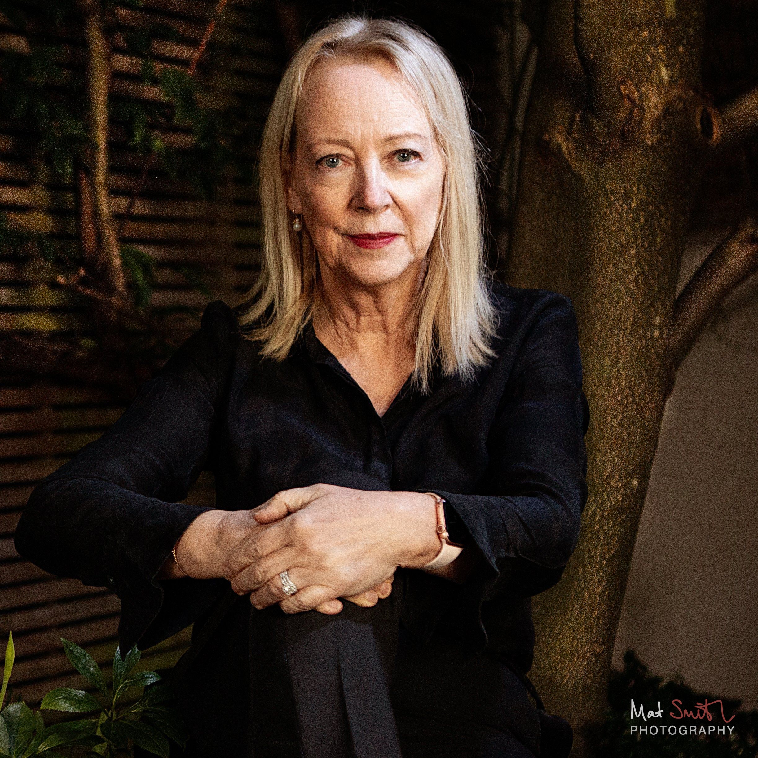 A woman with blonde hair in a black shirt leans forward with clasped hands beside a tree trunk in an evening garden, portrait by Mat Smith Photography, London