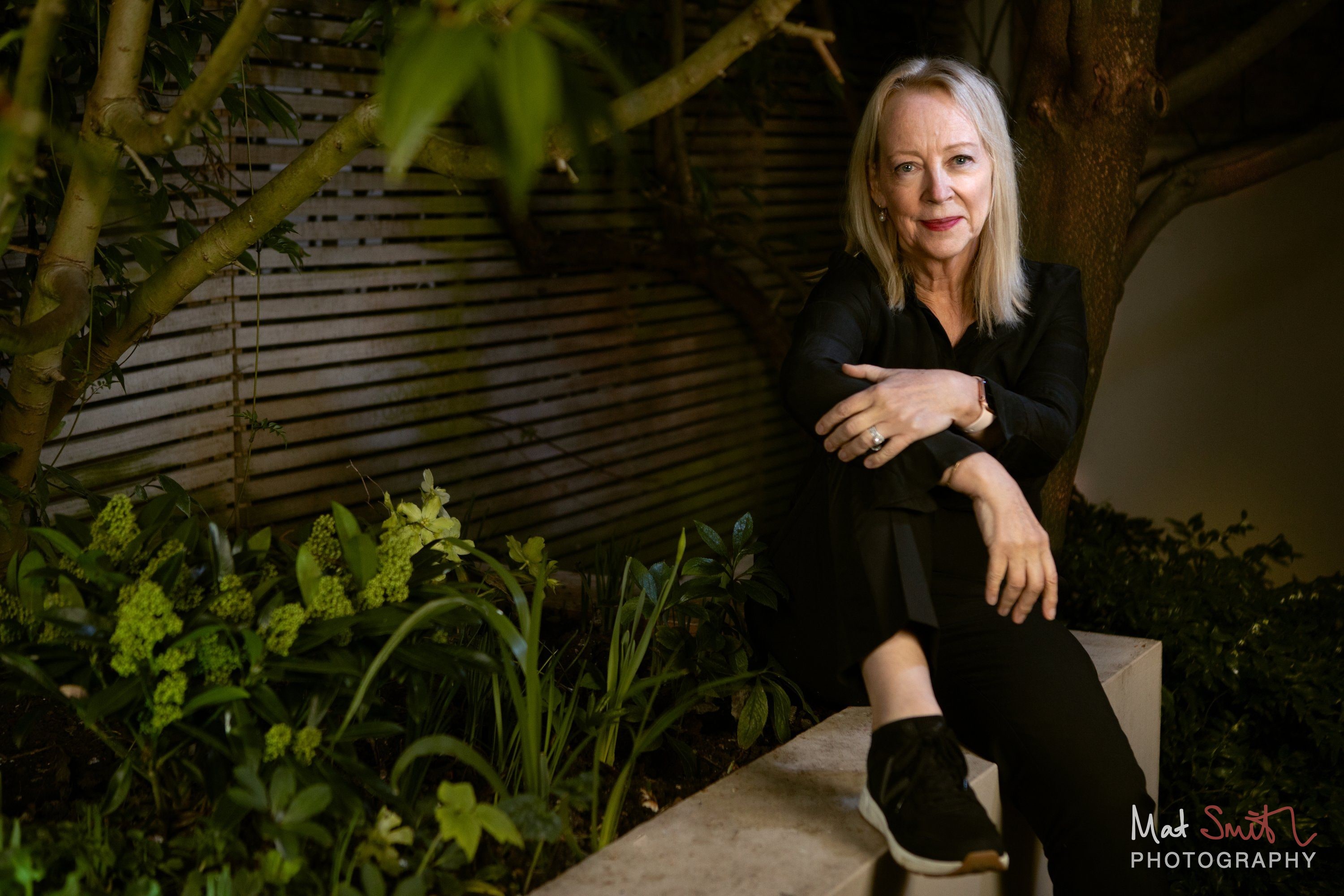 A woman in black sits on a stone garden wall with arms folded over her knee, surrounded by green leaves and flowers, portrait by Mat Smith Photography, London