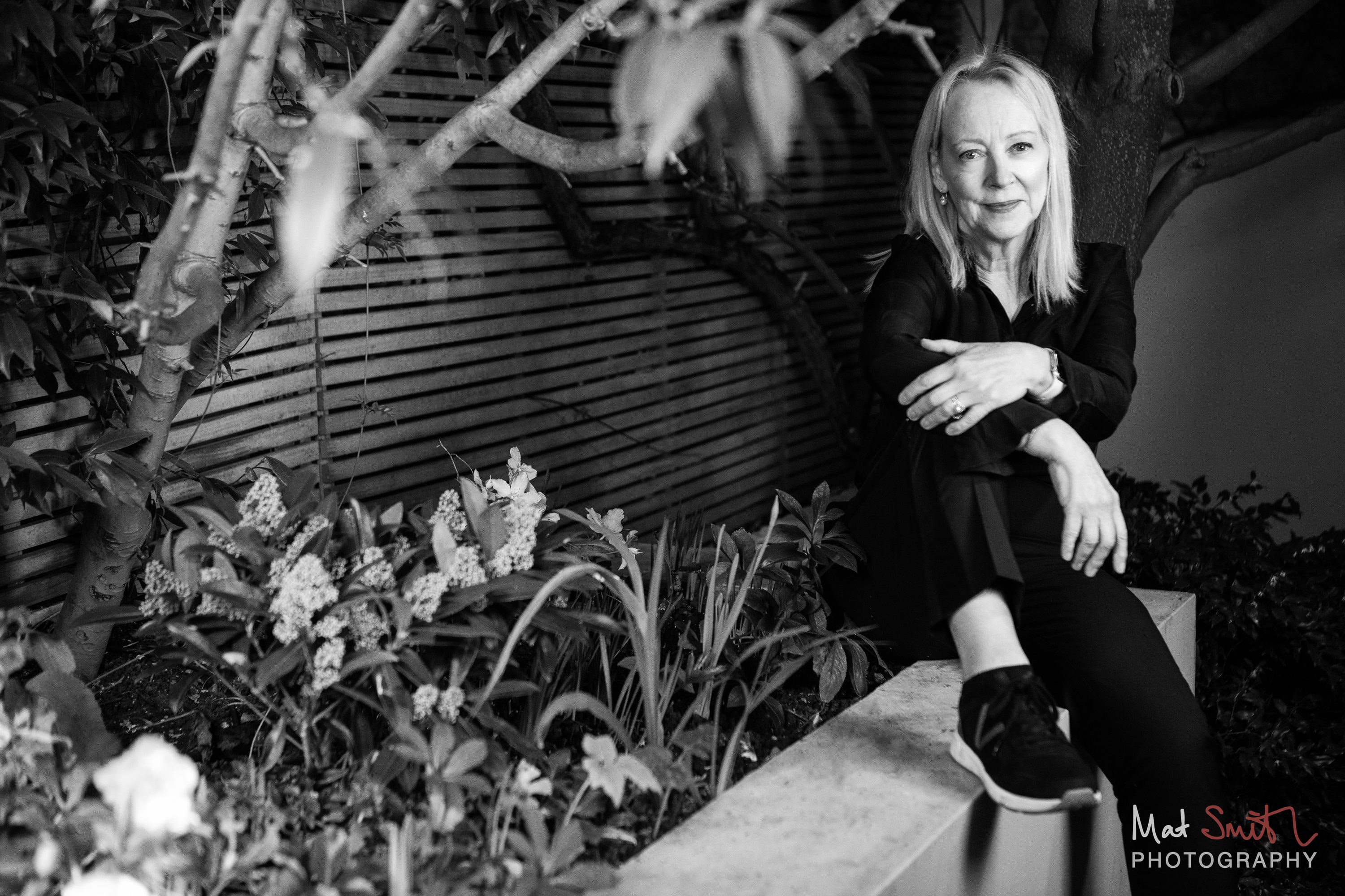 Black and white portrait of a woman seated on a garden wall framed by branches and pale flowers, portrait by Mat Smith Photography, London
