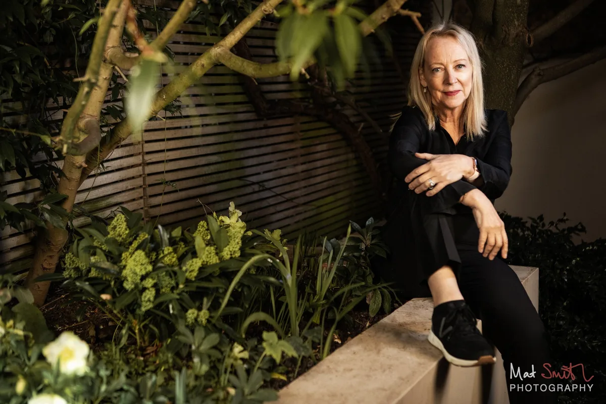 A woman in black seated on a low wall in a lit evening garden, with branches and spring flowers in the foreground, portrait by Mat Smith Photography, London