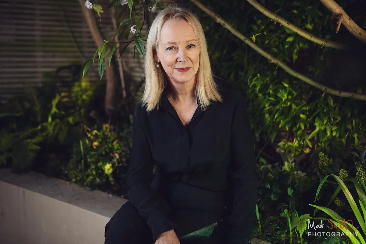 A woman with blonde hair in a black shirt sits on a pale garden wall in front of dense green foliage at night, portrait by Mat Smith Photography, London
