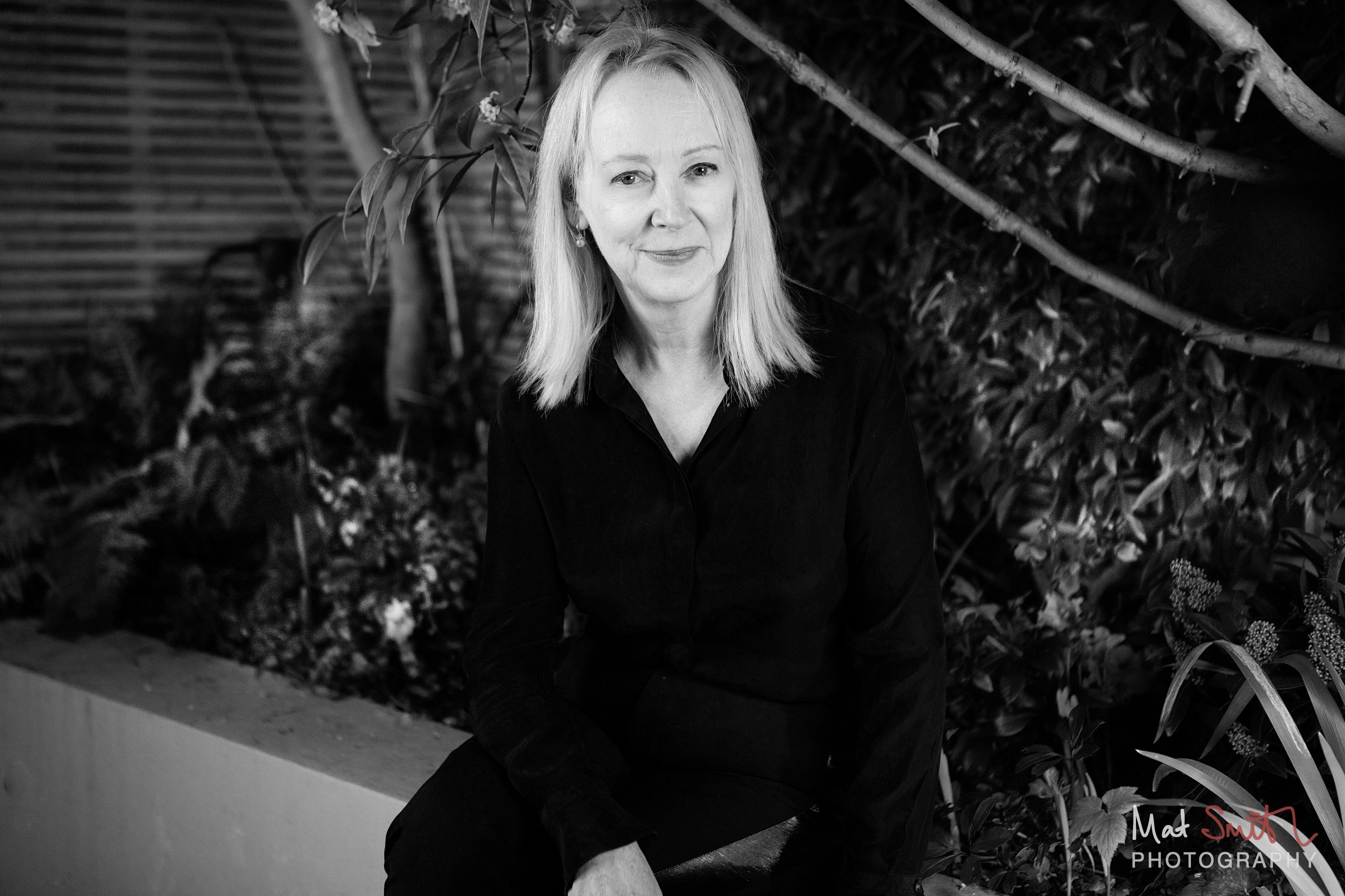 Black and white portrait of a woman in a black shirt seated on a garden wall against dark foliage, portrait by Mat Smith Photography, London