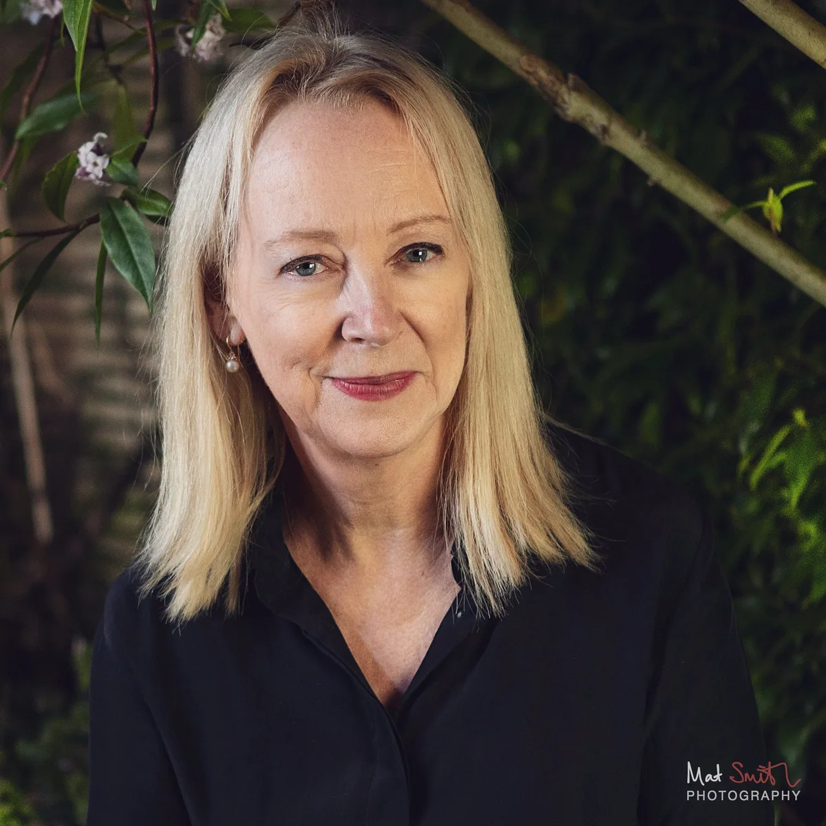 Head and shoulders portrait of a smiling woman with blonde hair in a black shirt among leaves and blossom, portrait by Mat Smith Photography, London