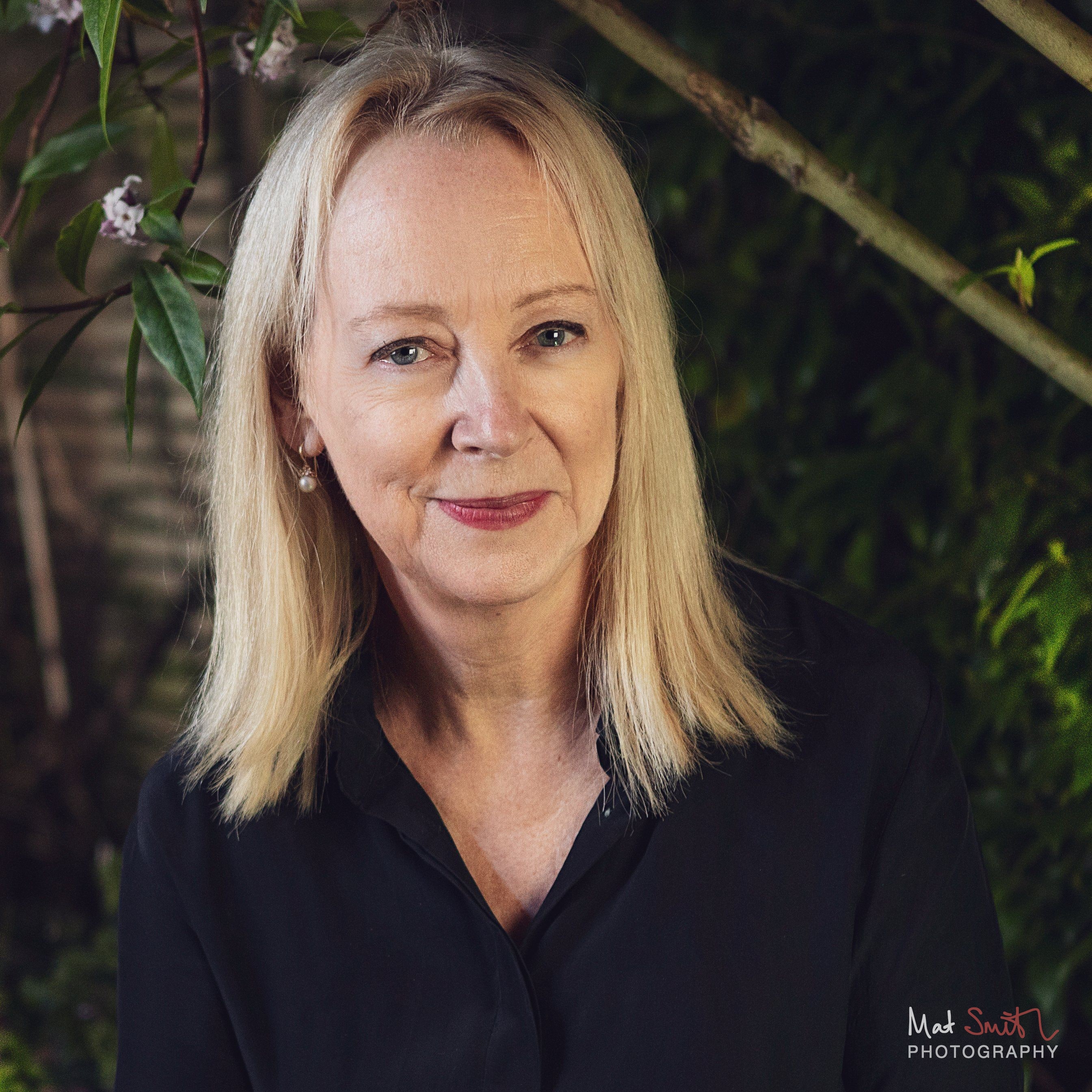 Head and shoulders portrait of a smiling woman with blonde hair in a black shirt among leaves and blossom, portrait by Mat Smith Photography, London