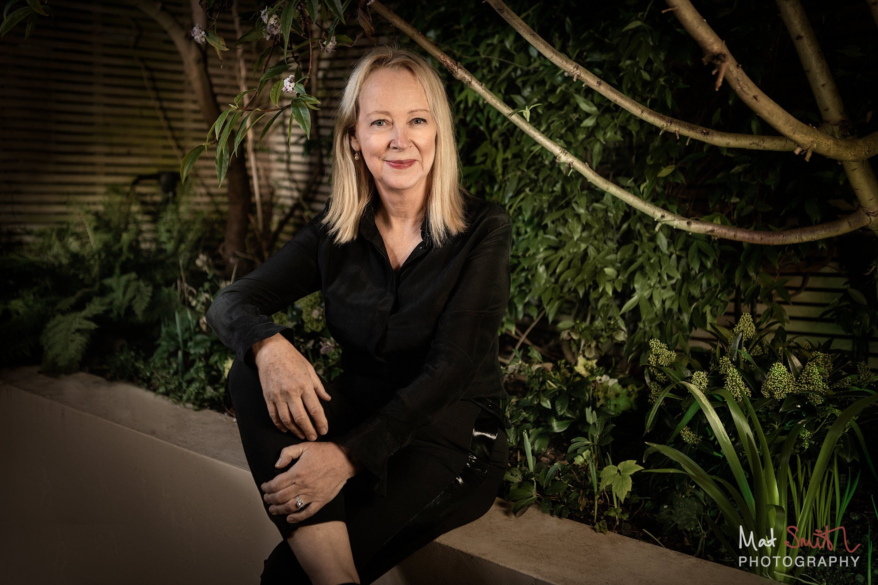 A woman in black sits on a garden wall with one hand on her knee under arching branches in warm evening light, portrait by Mat Smith Photography, London