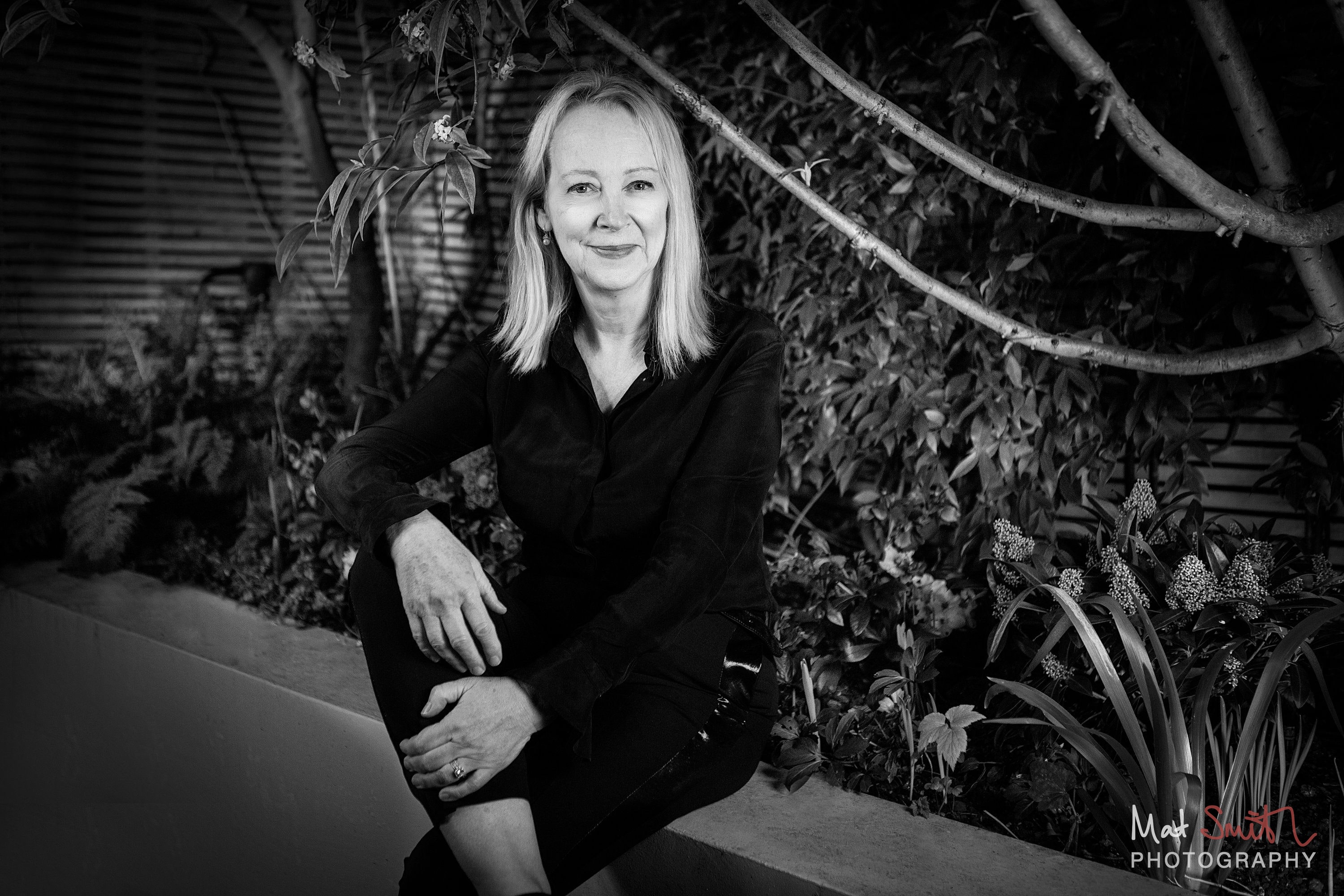 Black and white portrait of a woman seated on a garden wall under arching branches, one hand resting on her knee, portrait by Mat Smith Photography, London