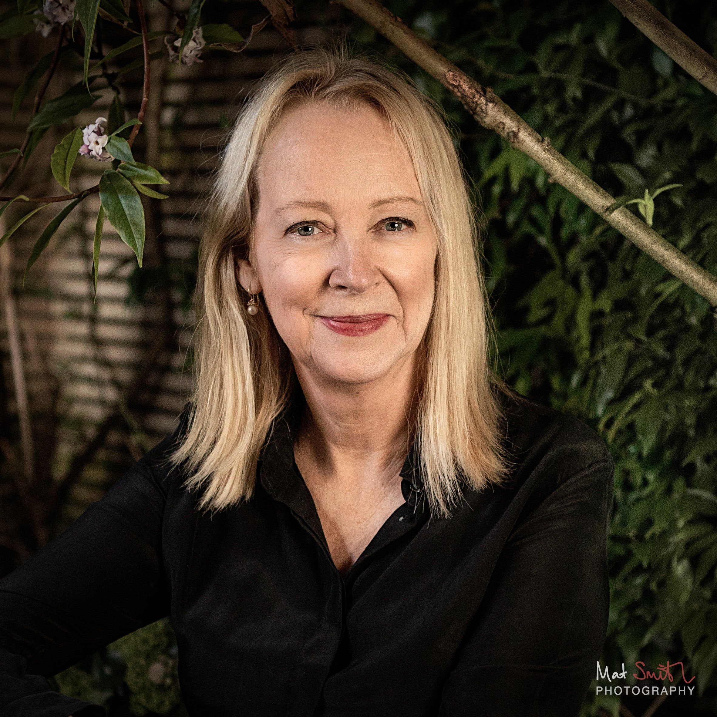 Close portrait of a woman with blonde hair smiling softly with blossom and branches around her in the dark garden, portrait by Mat Smith Photography, London