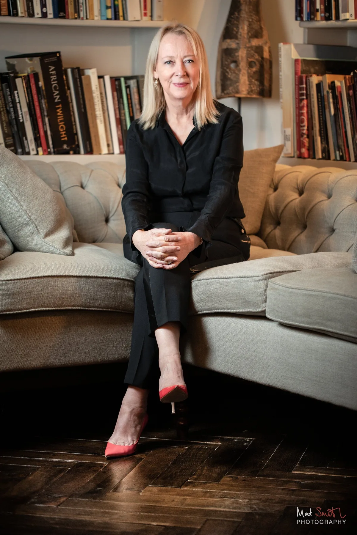 A woman in a black outfit and red heeled shoes sits on a pale buttoned sofa in front of full bookshelves, portrait by Mat Smith Photography, London