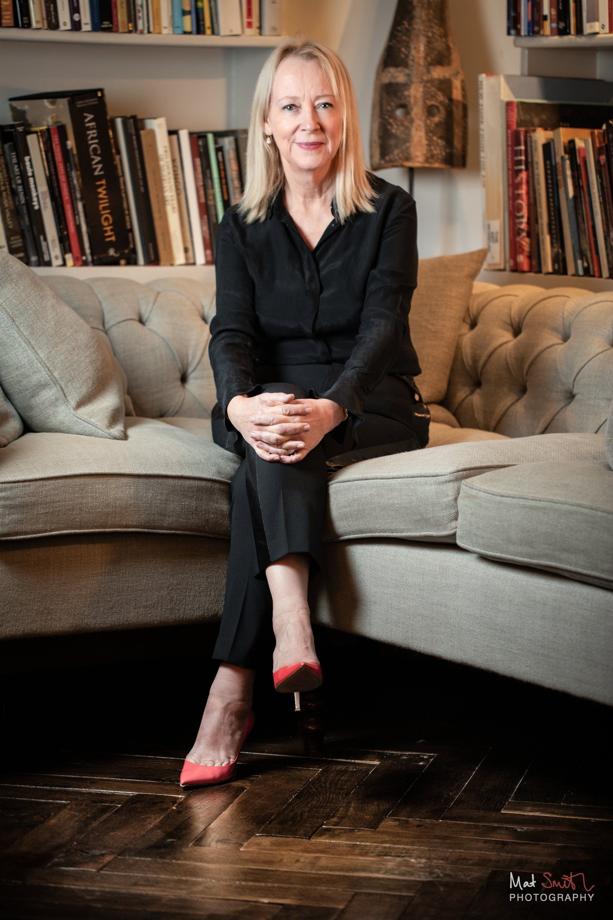 A woman in a black outfit and red heeled shoes sits on a pale buttoned sofa in front of full bookshelves, portrait by Mat Smith Photography, London