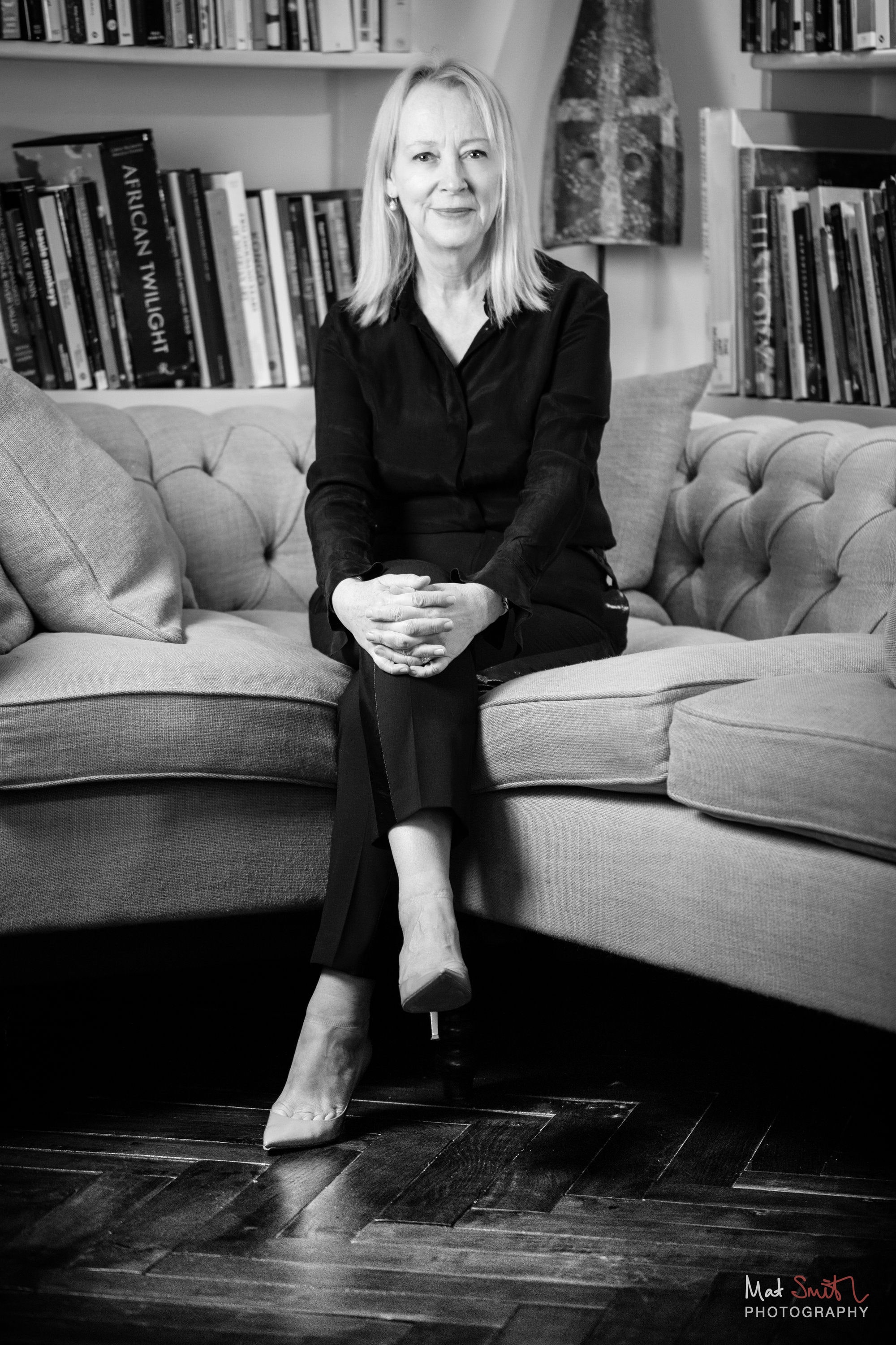Black and white portrait of a woman in black seated on a buttoned sofa in front of bookshelves, legs crossed at the ankle, portrait by Mat Smith Photography, London