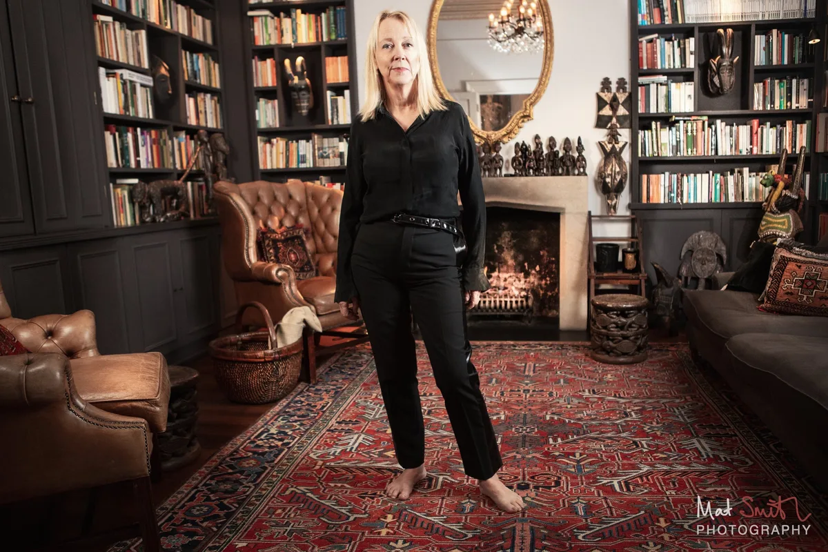 A barefoot woman in black stands on a red patterned rug in a book-lined sitting room with a lit fireplace and leather armchairs, portrait by Mat Smith Photography, London
