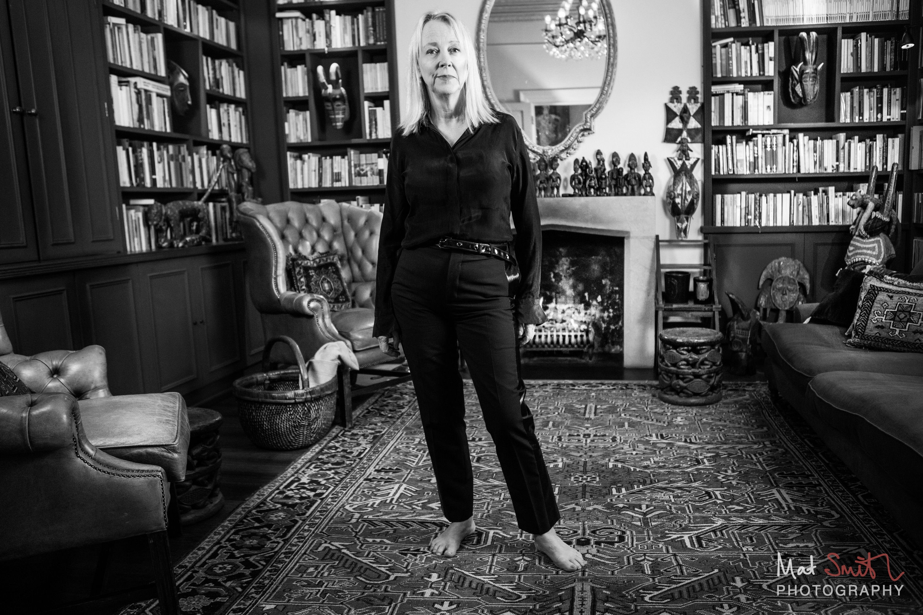 Black and white view of a barefoot woman in black standing in a book-lined sitting room with a fire, mirror and armchairs, portrait by Mat Smith Photography, London