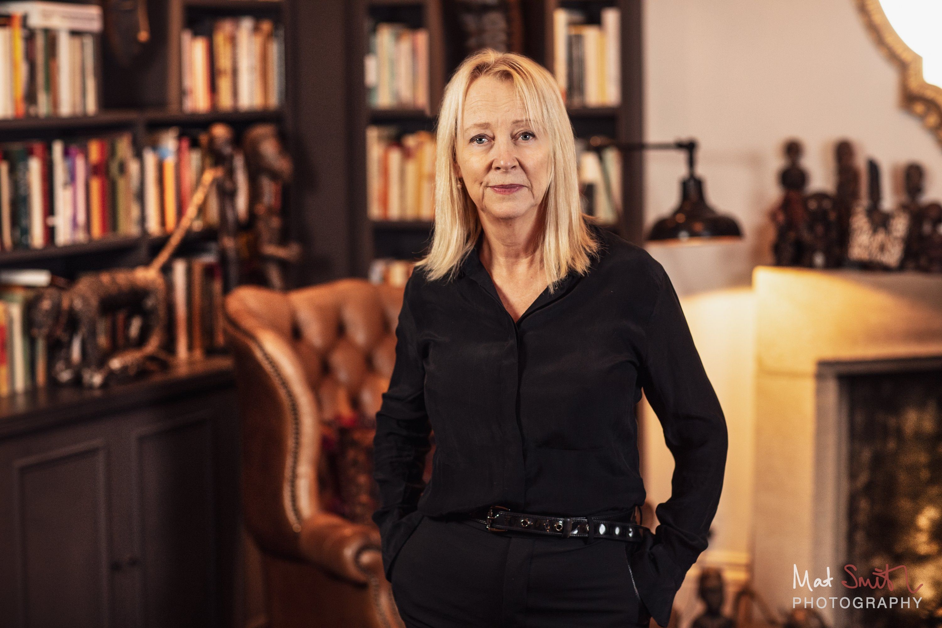 A woman with blonde hair in a black shirt stands with hands in pockets beside a leather wing chair and bookshelves, portrait by Mat Smith Photography, London
