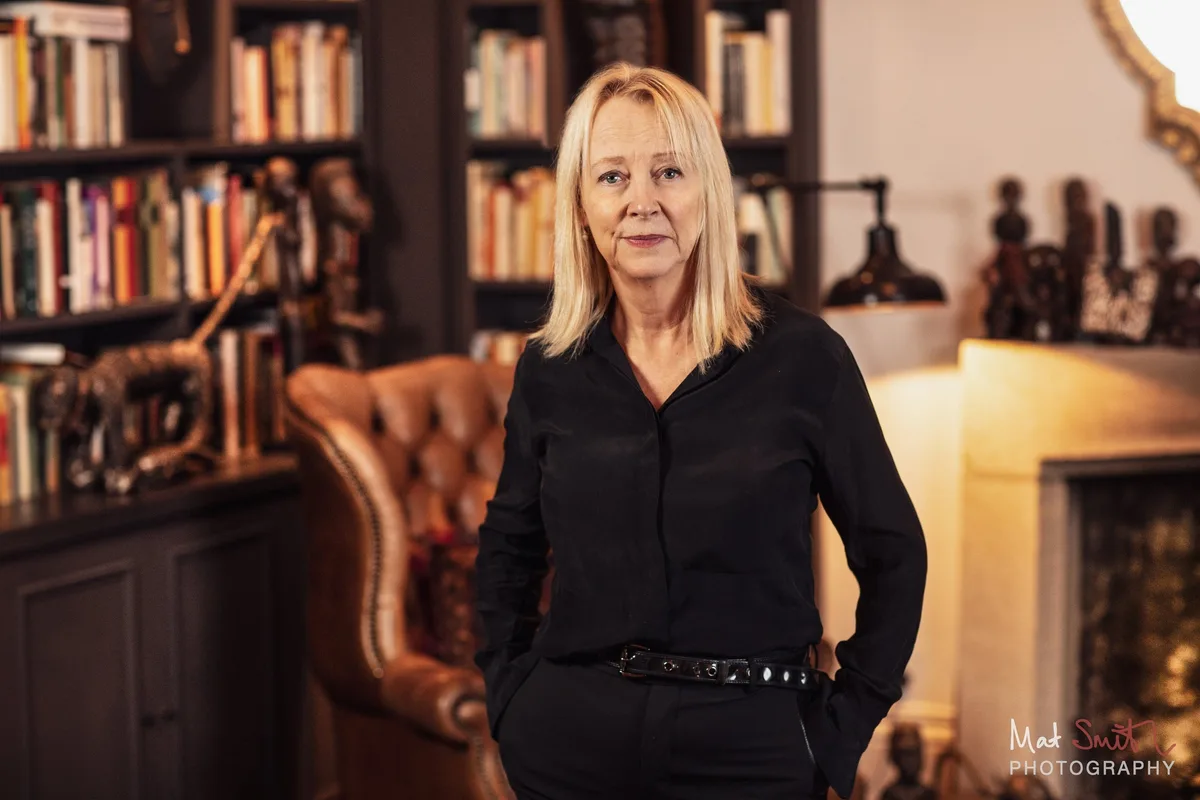 A woman with blonde hair in a black shirt stands with hands in pockets beside a leather wing chair and bookshelves, portrait by Mat Smith Photography, London