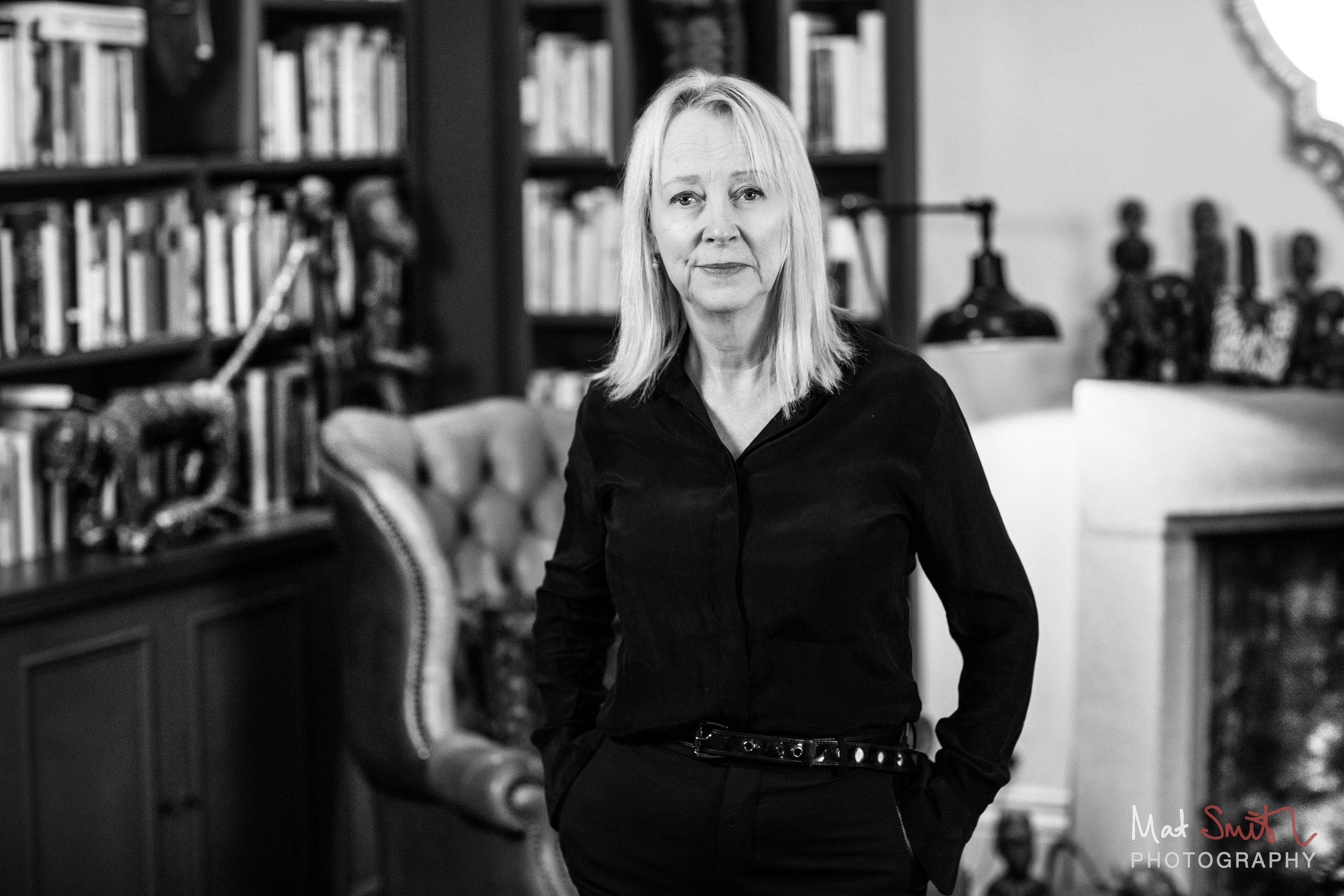Black and white portrait of a woman in a black shirt with hands in her pockets beside a wing chair and bookshelves, portrait by Mat Smith Photography, London