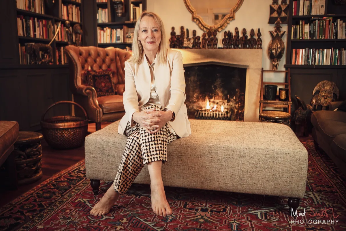 A barefoot woman in a cream jacket and checked trousers sits on a large ottoman in front of a lit fireplace and bookshelves, portrait by Mat Smith Photography, London