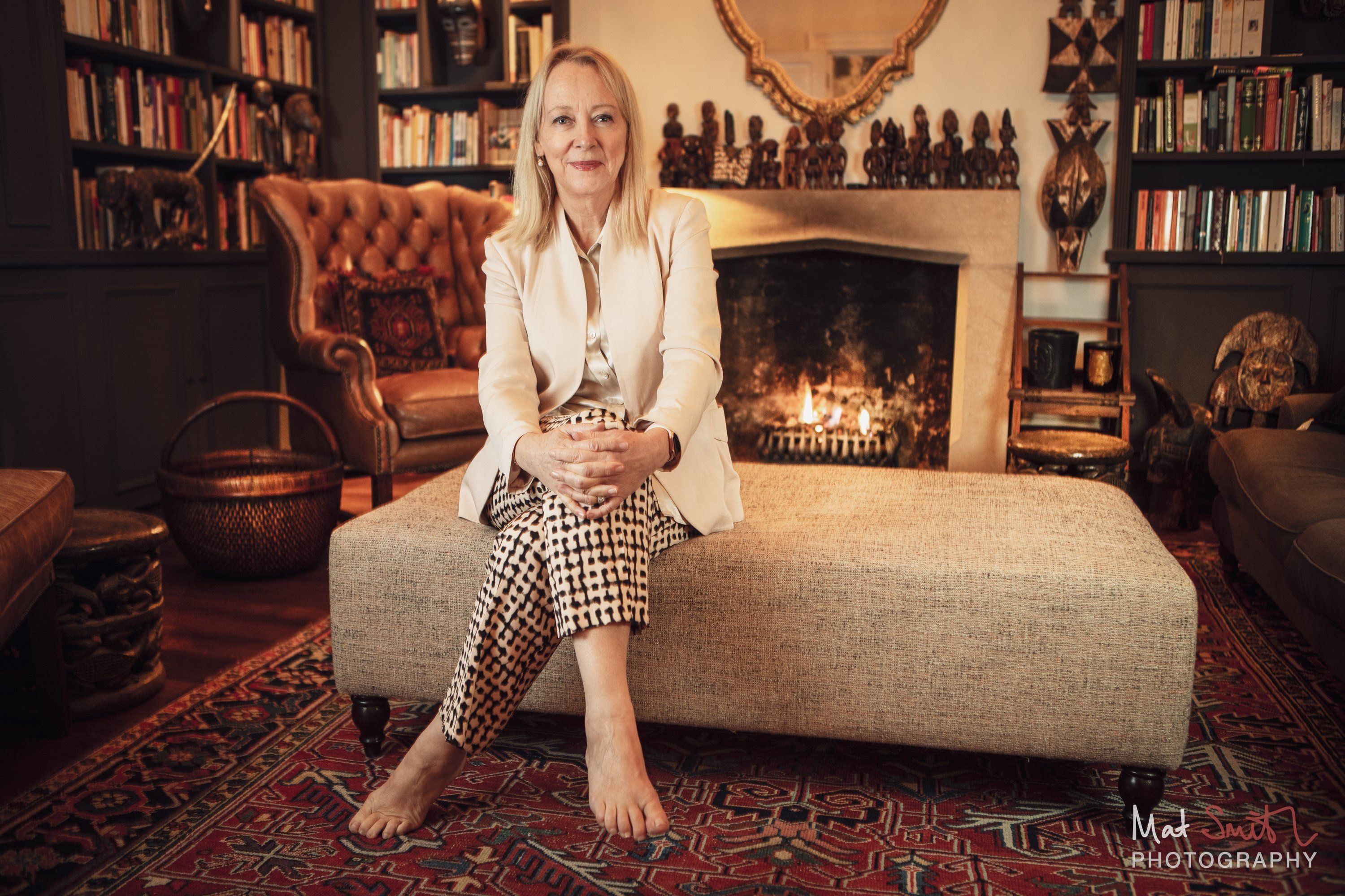 A barefoot woman in a cream jacket and checked trousers sits on a large ottoman in front of a lit fireplace and bookshelves, portrait by Mat Smith Photography, London