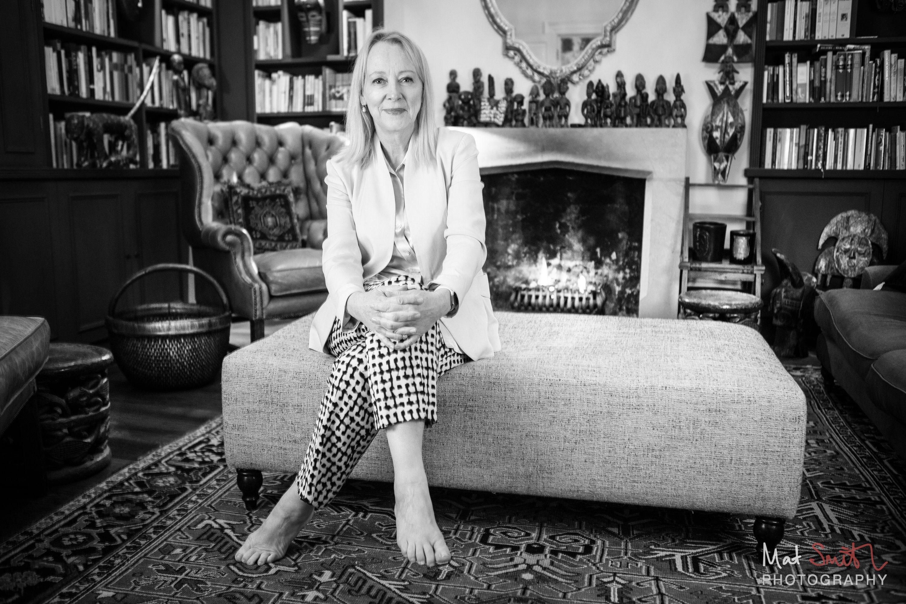 Black and white portrait of a woman in a pale jacket and checked trousers seated on an ottoman before an open fire, portrait by Mat Smith Photography, London