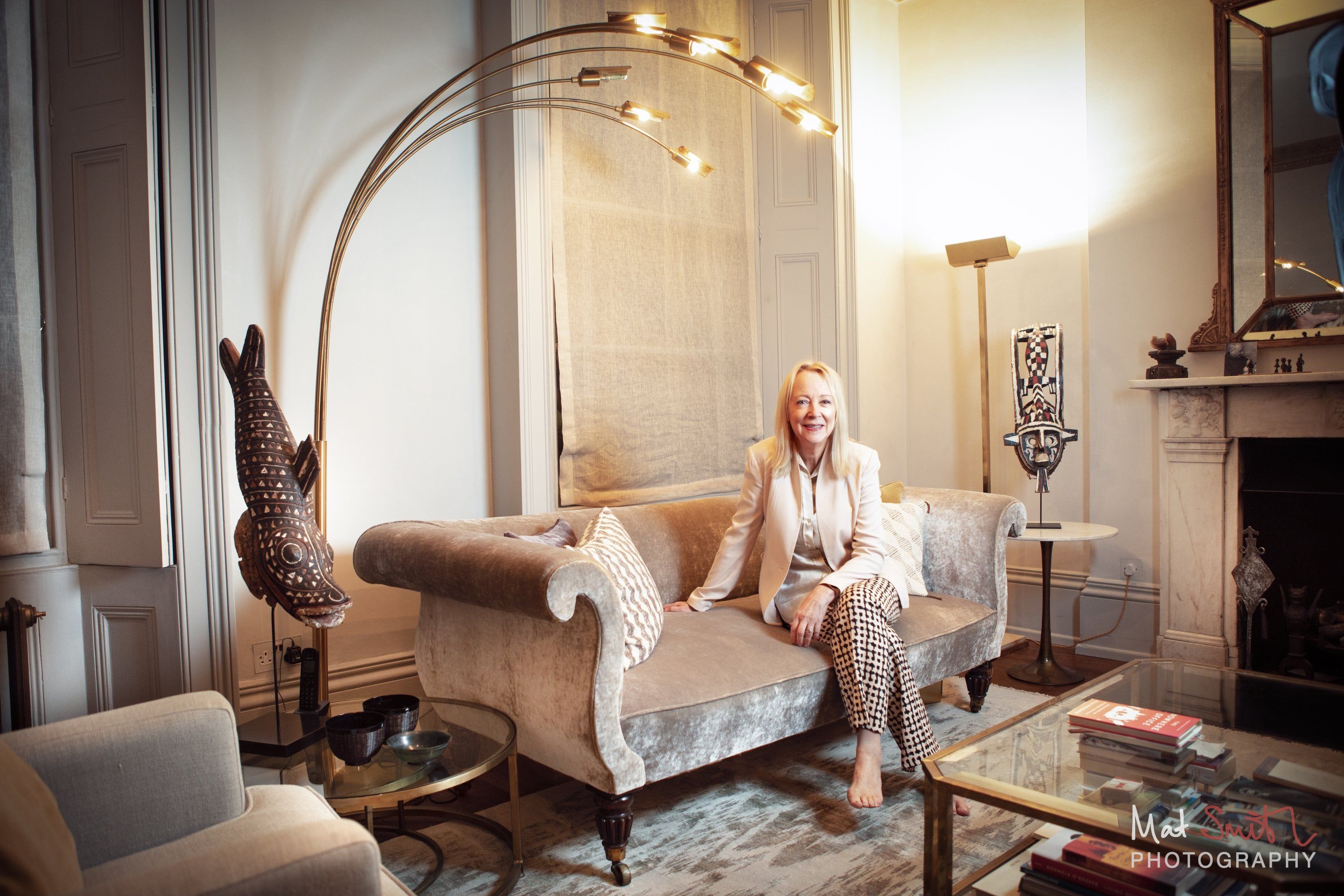 Wide view of a woman in a cream jacket seated on a velvet sofa in a tall drawing room with an arc lamp and marble fireplace, portrait by Mat Smith Photography, London