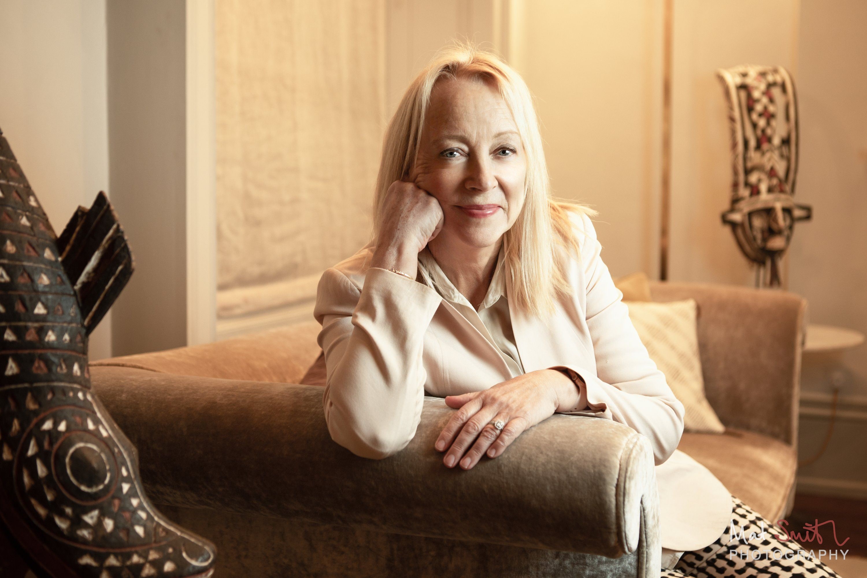 A woman in a cream jacket leans on the arm of a sofa with her chin on her hand in soft warm window light, portrait by Mat Smith Photography, London