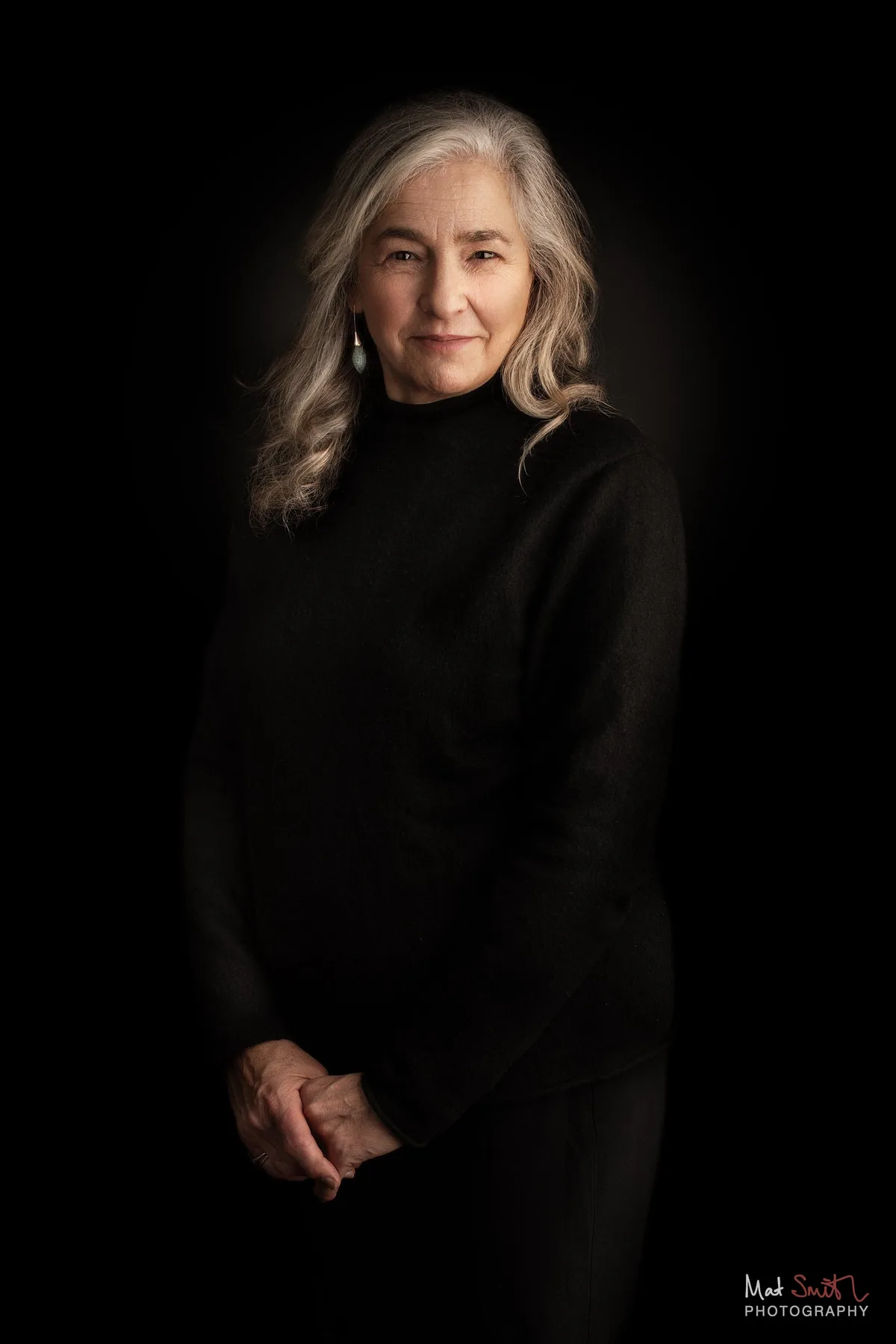 A woman with long wavy grey hair in a black roll neck stands with hands clasped against a black backdrop, portrait by Mat Smith Photography, London