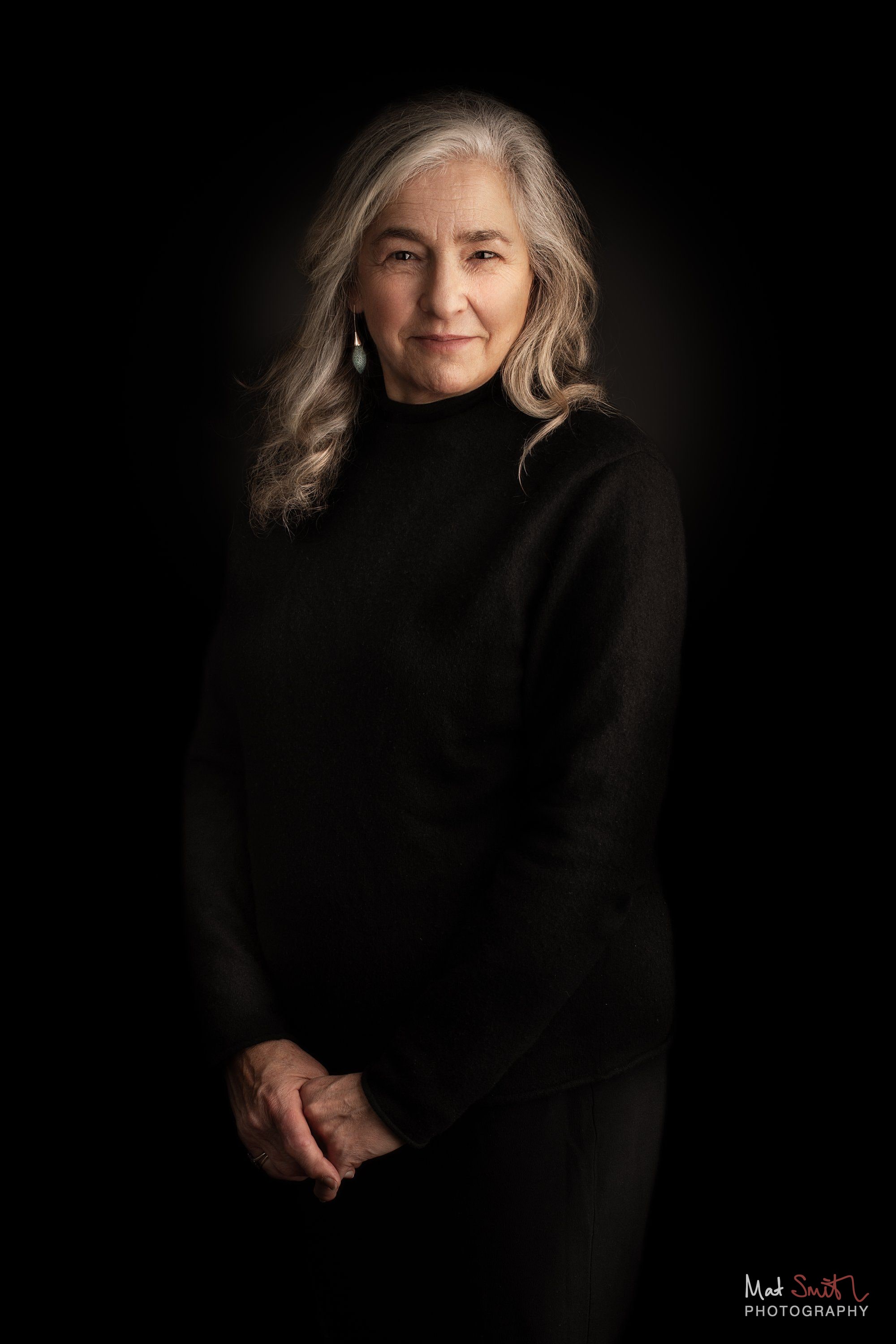 A woman with long wavy grey hair in a black roll neck stands with hands clasped against a black backdrop, portrait by Mat Smith Photography, London