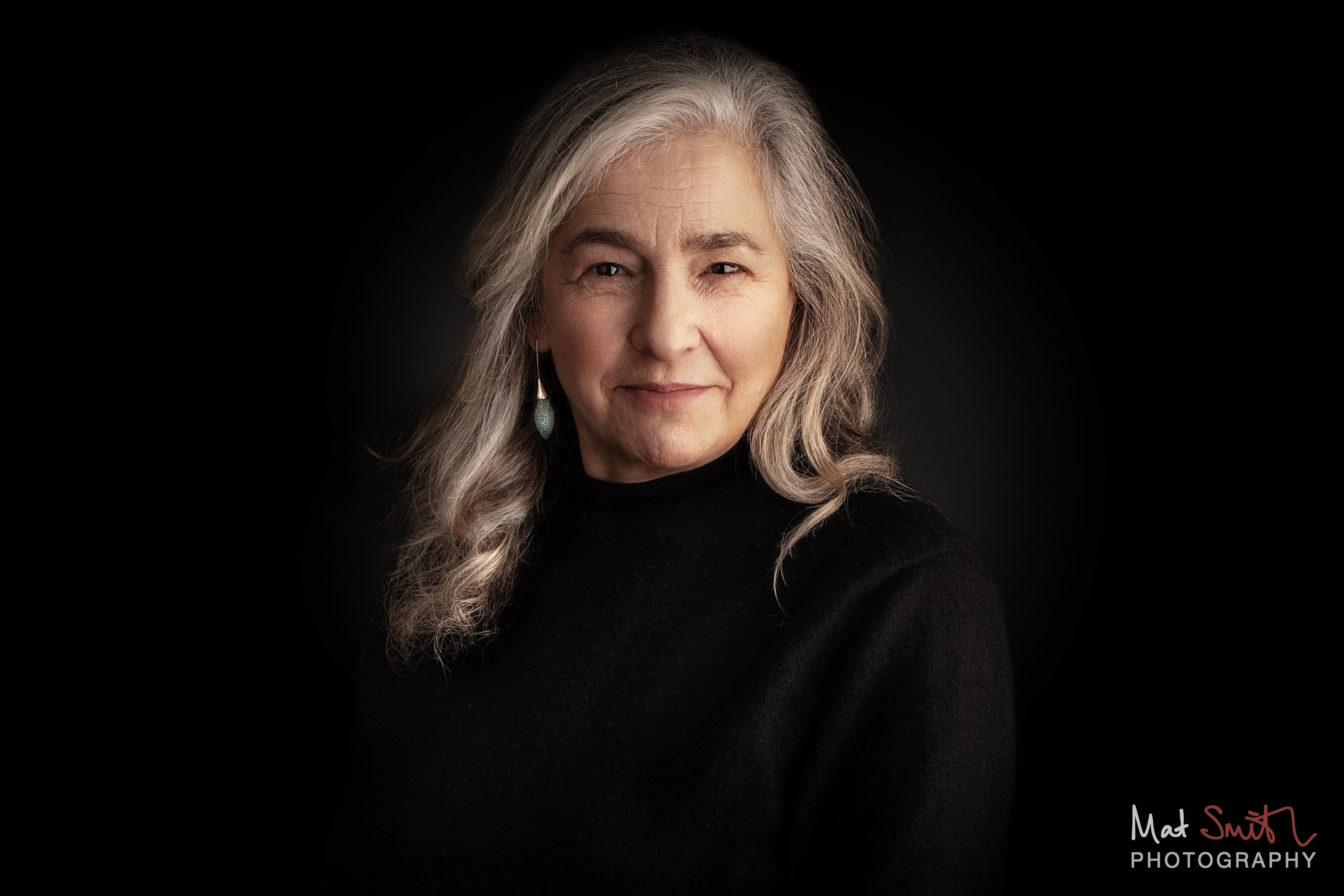 Head and shoulders portrait of a woman with long wavy grey hair and drop earrings in a black roll neck against black, portrait by Mat Smith Photography, London