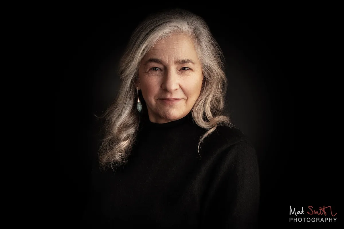 Head and shoulders portrait of a woman with long wavy grey hair and drop earrings in a black roll neck against black, portrait by Mat Smith Photography, London