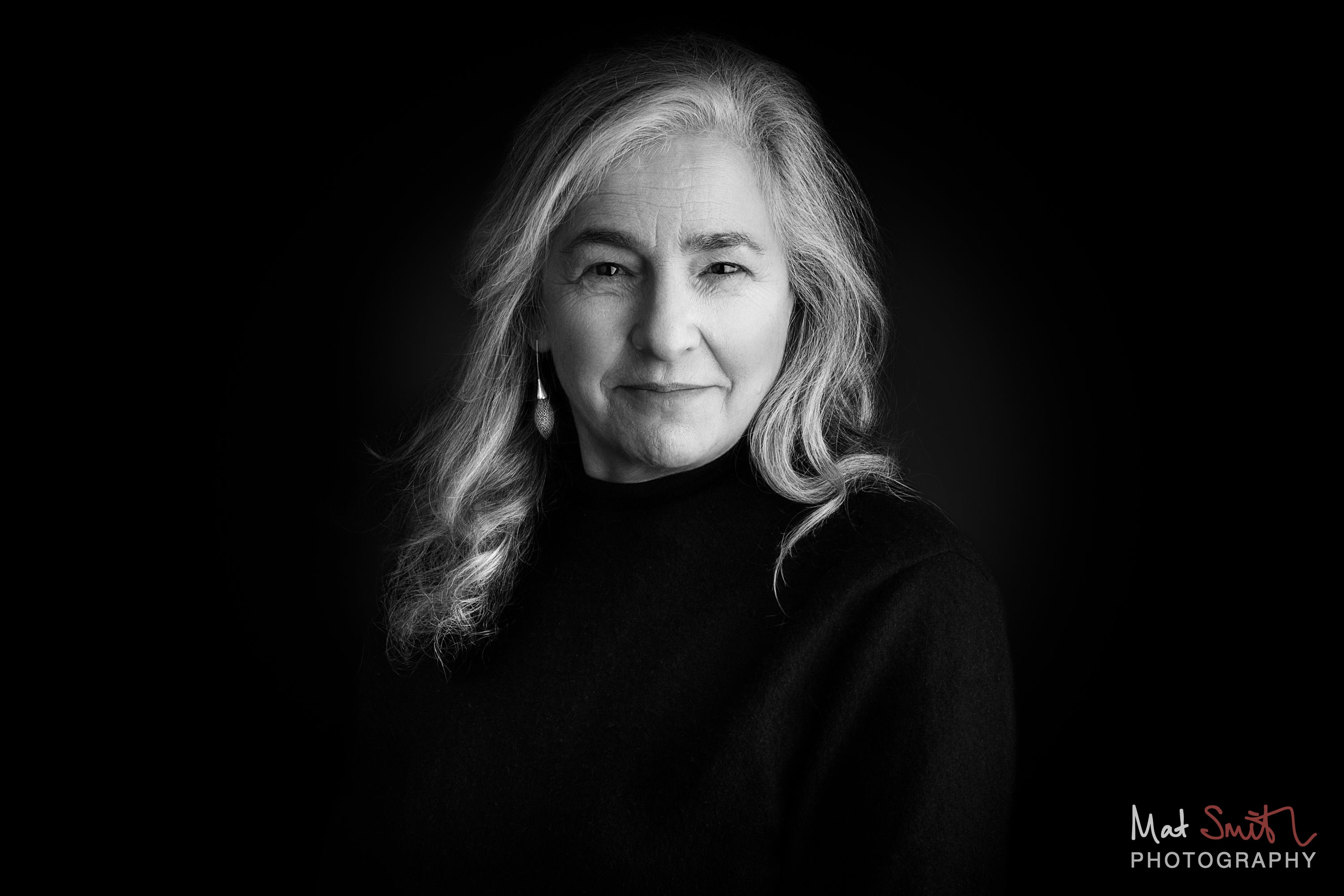 Black and white head and shoulders portrait of a woman with long wavy grey hair in a black roll neck against black, portrait by Mat Smith Photography, London