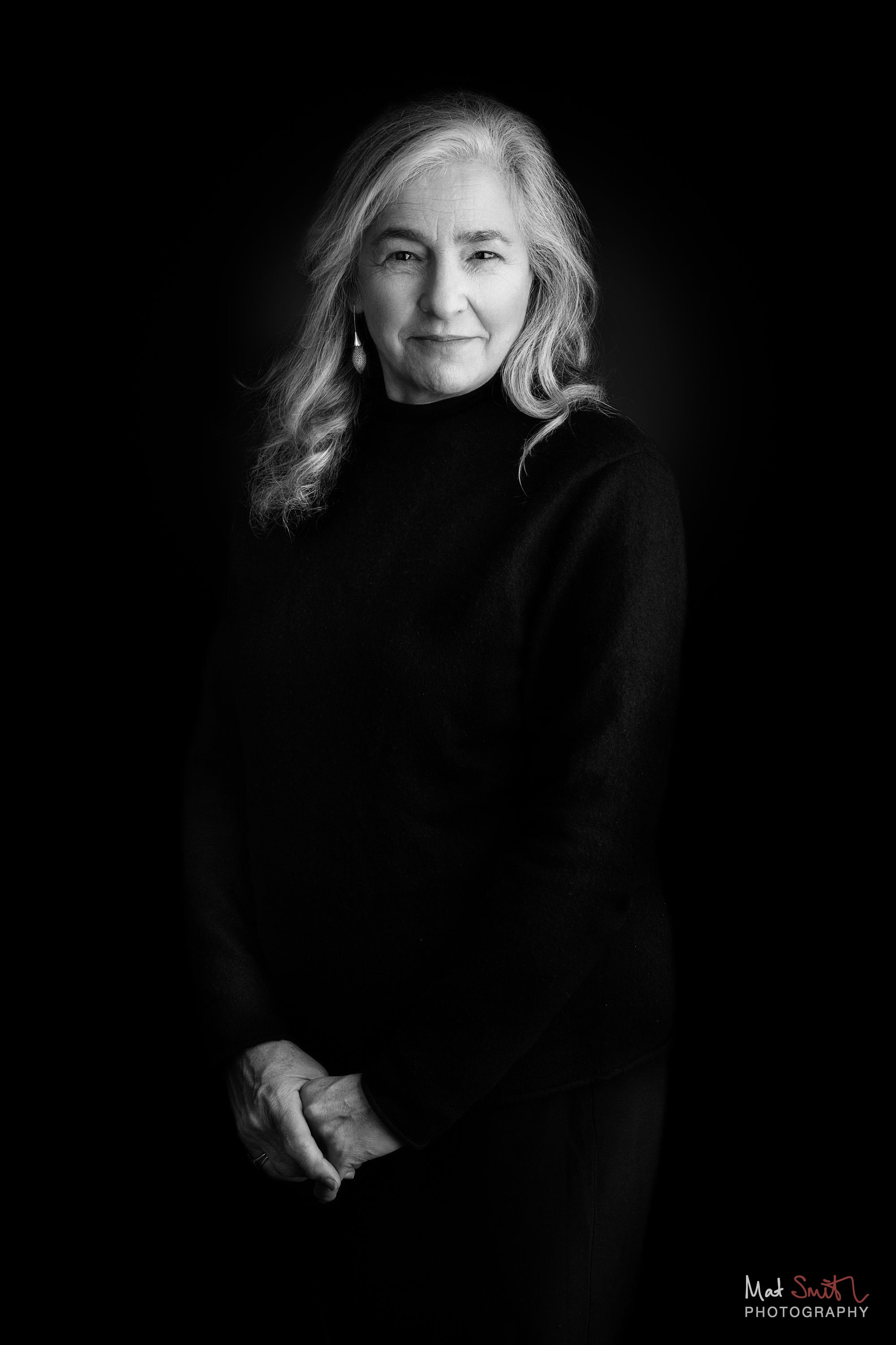 Black and white portrait of a woman with long grey hair standing with hands clasped against a black backdrop, portrait by Mat Smith Photography, London