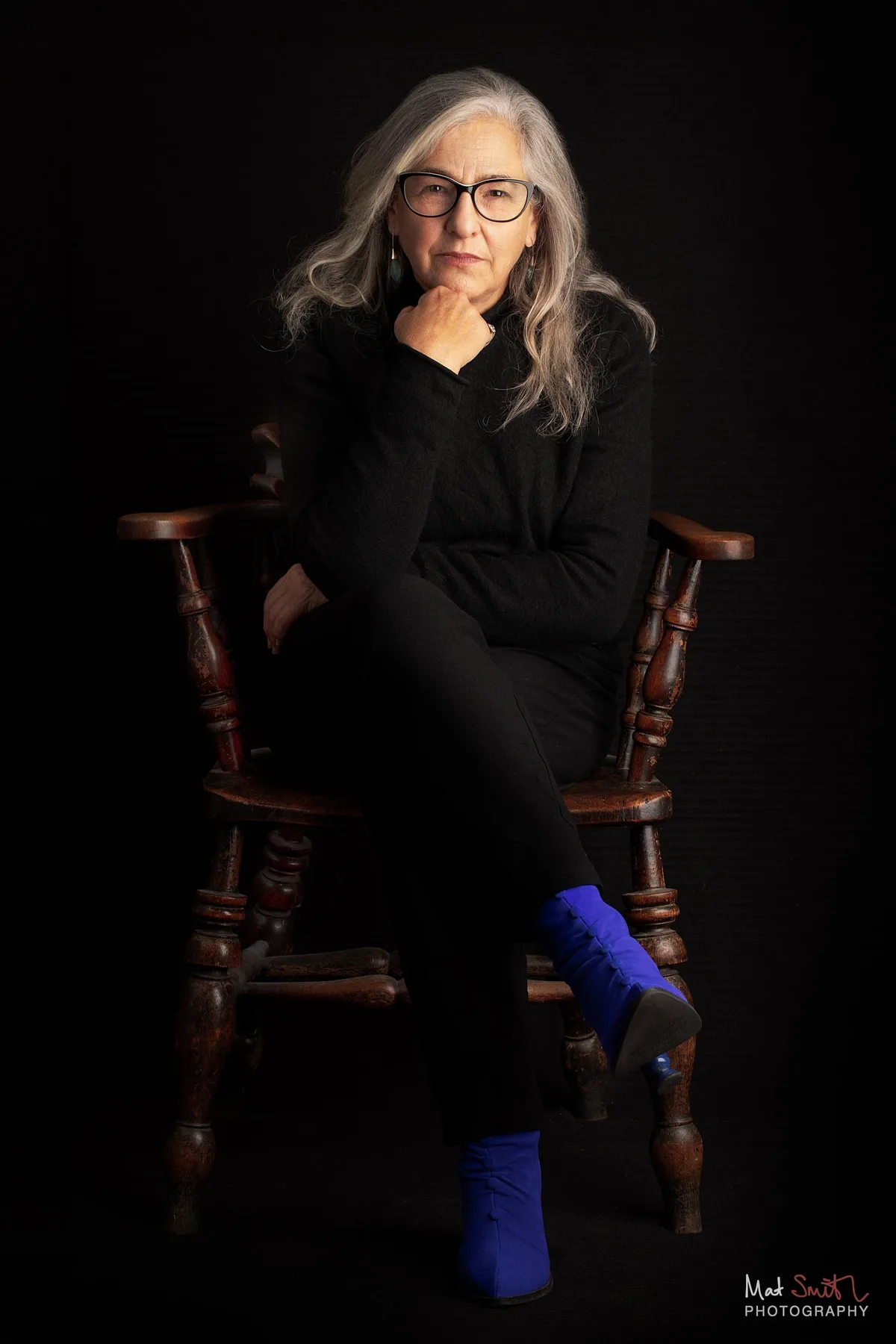A woman in glasses and black clothes sits cross-legged in a wooden spindle chair with her hand to her chin, wearing bright blue boots, portrait by Mat Smith Photography, London
