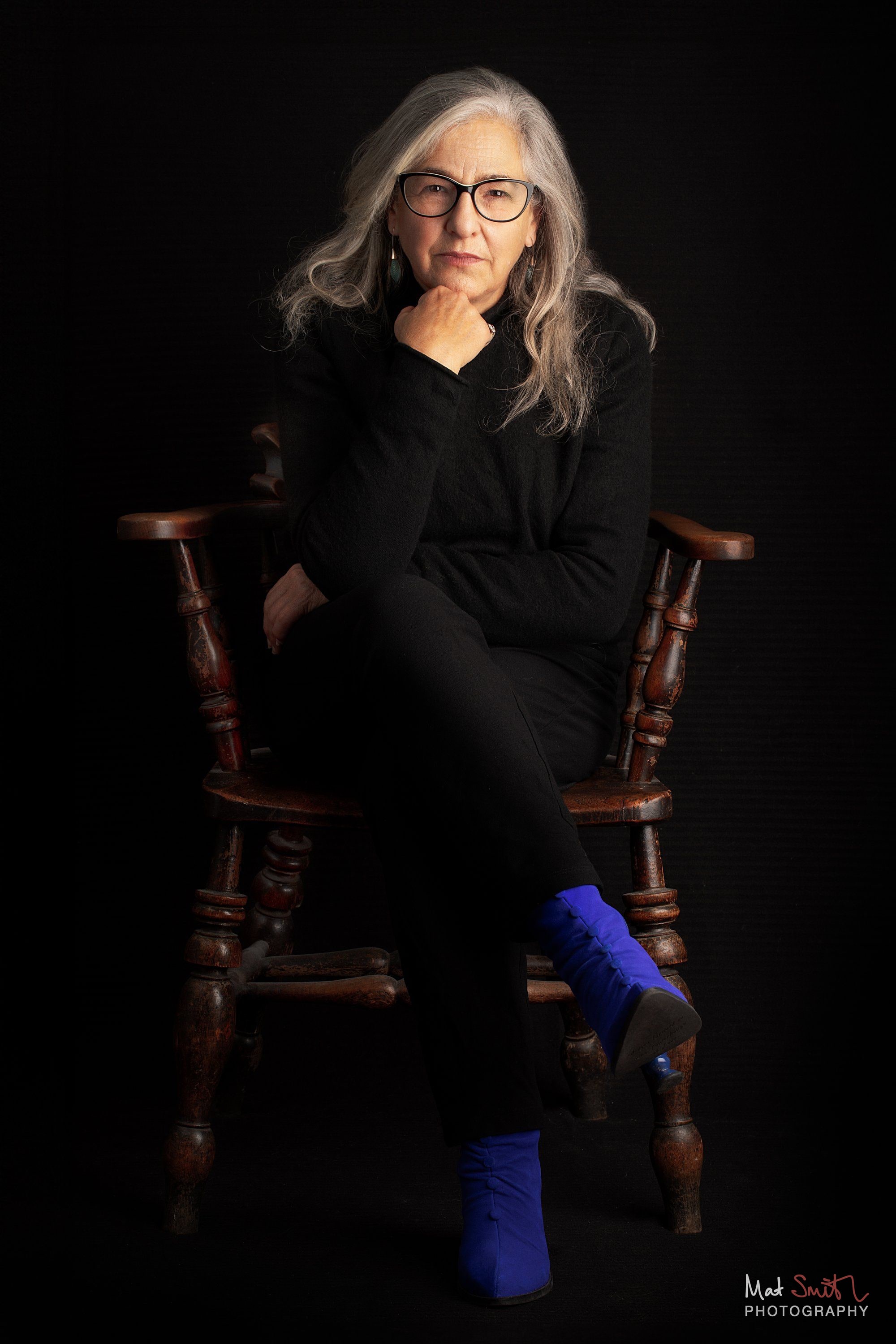 A woman in glasses and black clothes sits cross-legged in a wooden spindle chair with her hand to her chin, wearing bright blue boots, portrait by Mat Smith Photography, London