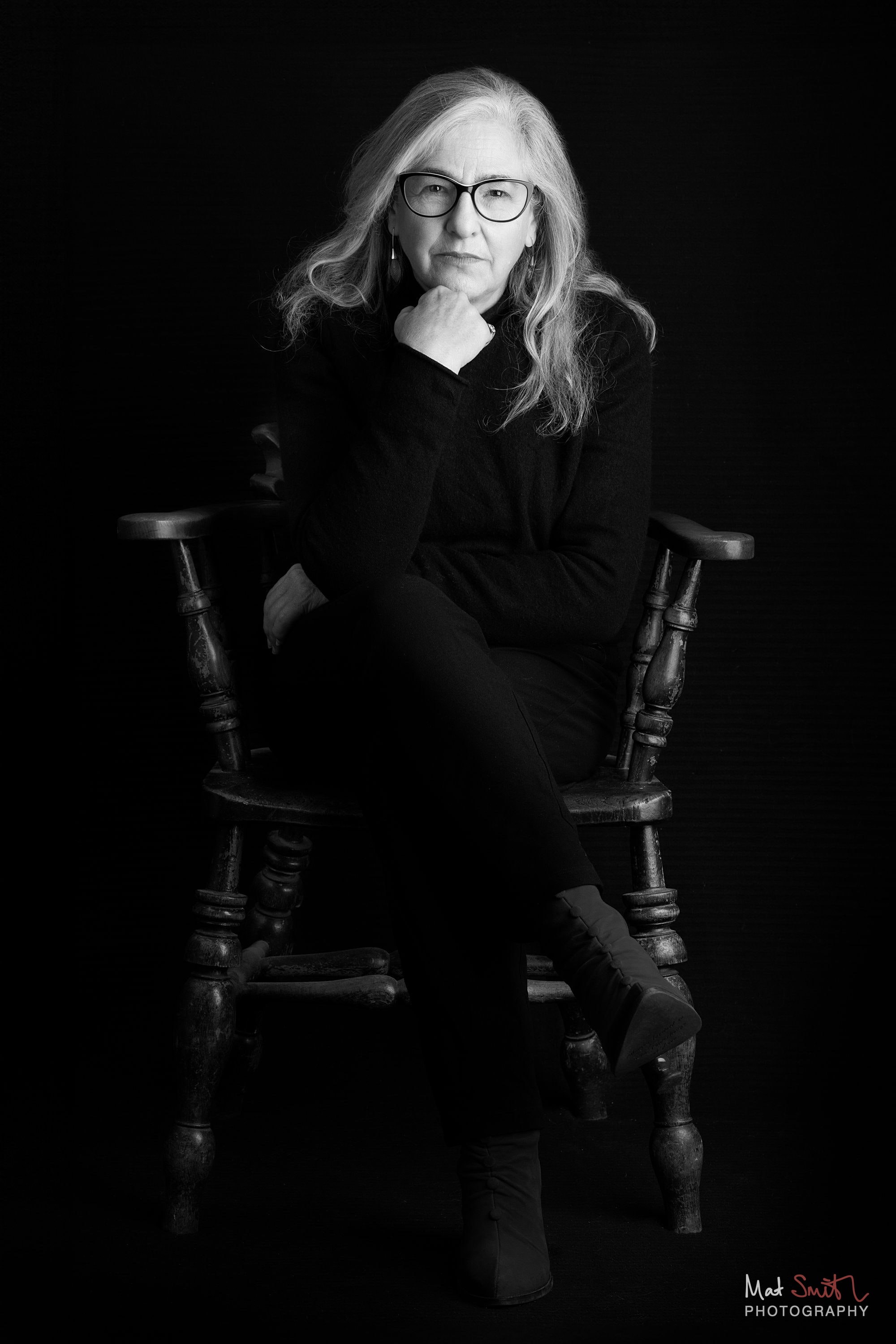 Black and white portrait of a woman in glasses seated cross-legged in a wooden spindle chair with her hand to her chin, portrait by Mat Smith Photography, London