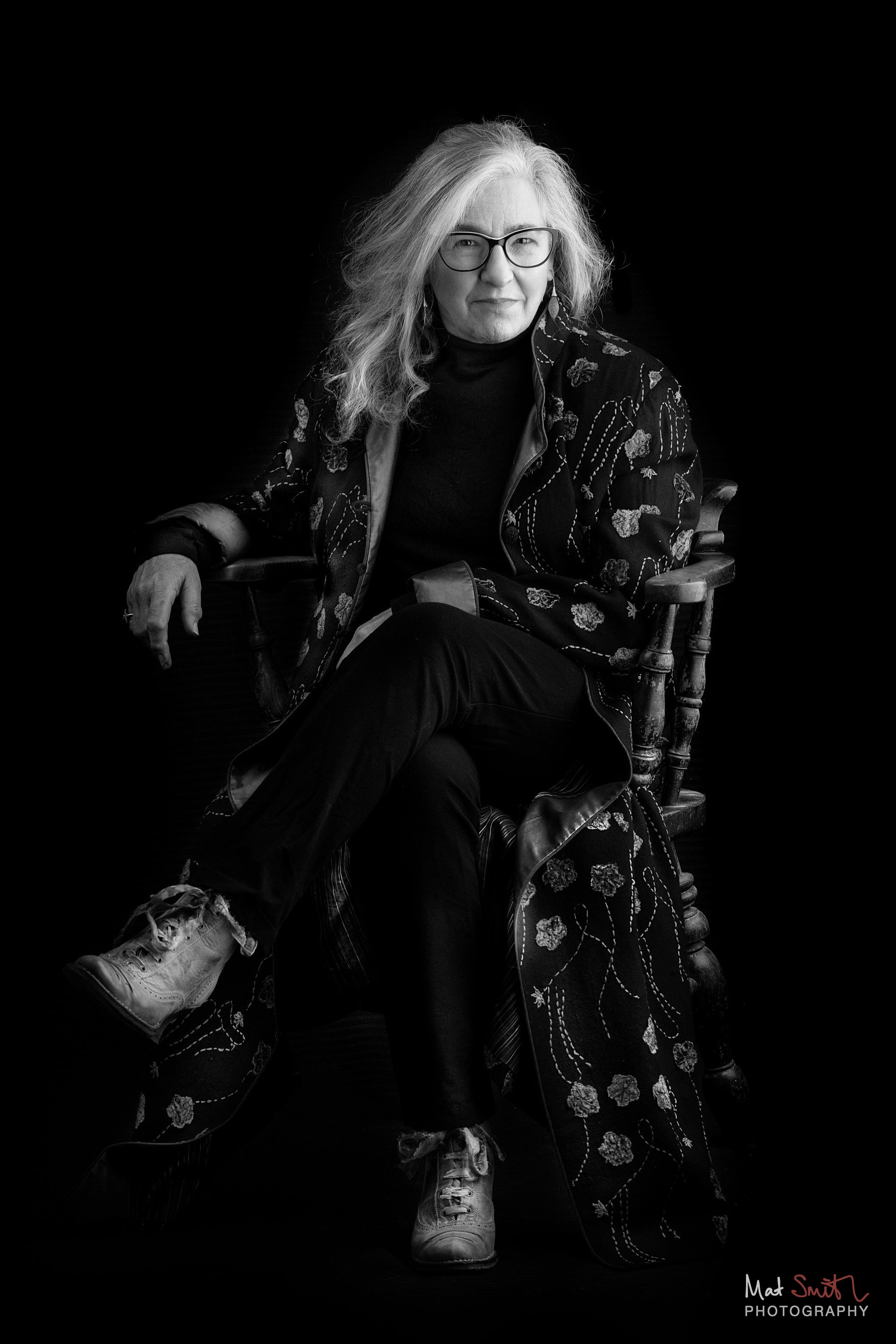 Black and white portrait of a woman in glasses and a long embroidered coat seated in a wooden chair against black, portrait by Mat Smith Photography, London